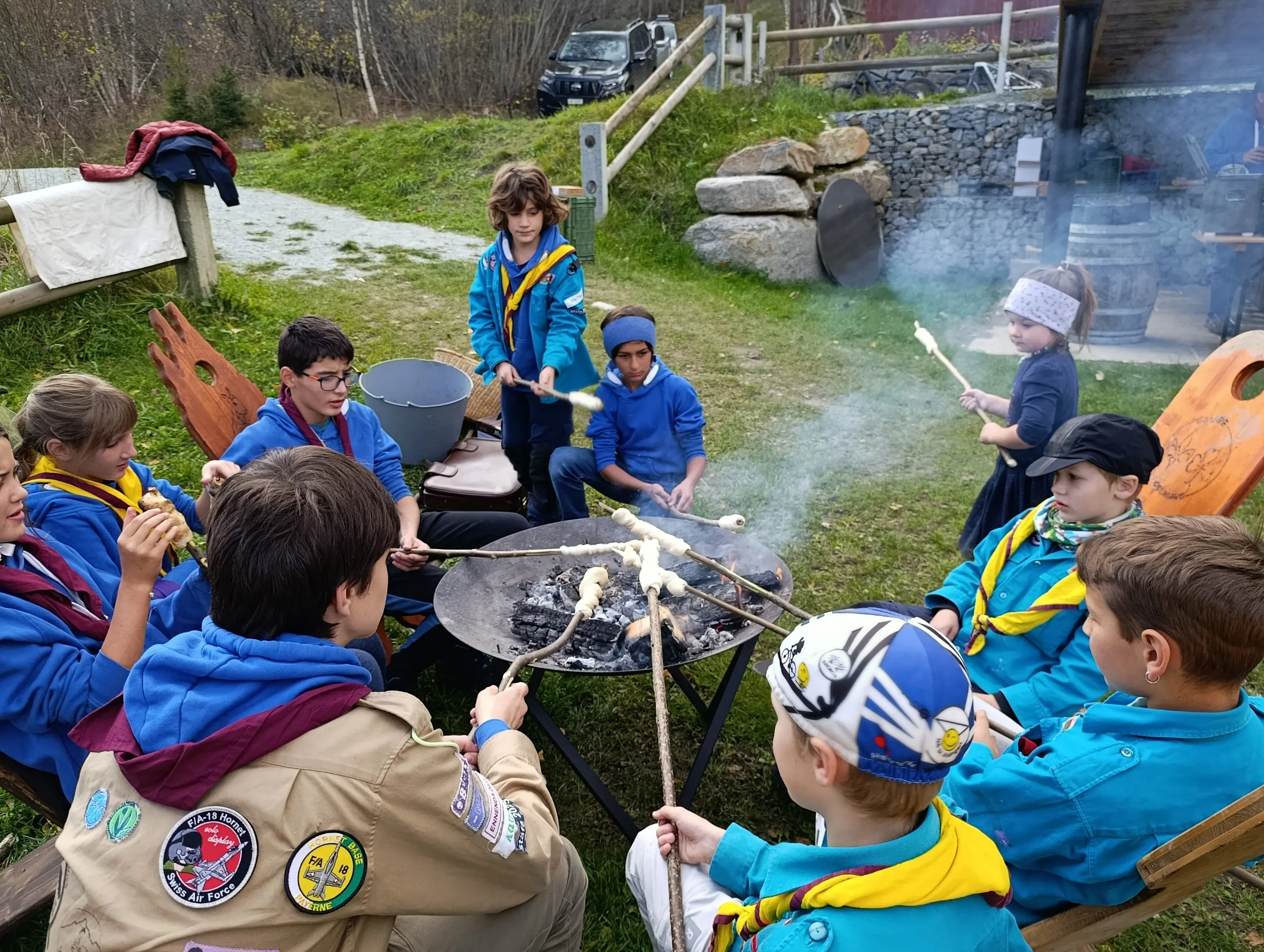 Un gruppo di ragazzi in abbigliamento scout si riunisce attorno a un fuoco all'aperto, mentre arrostiscono del pane su bastoni. Sullo sfondo ci sono attrezzature per cucinare e un paesaggio naturale.