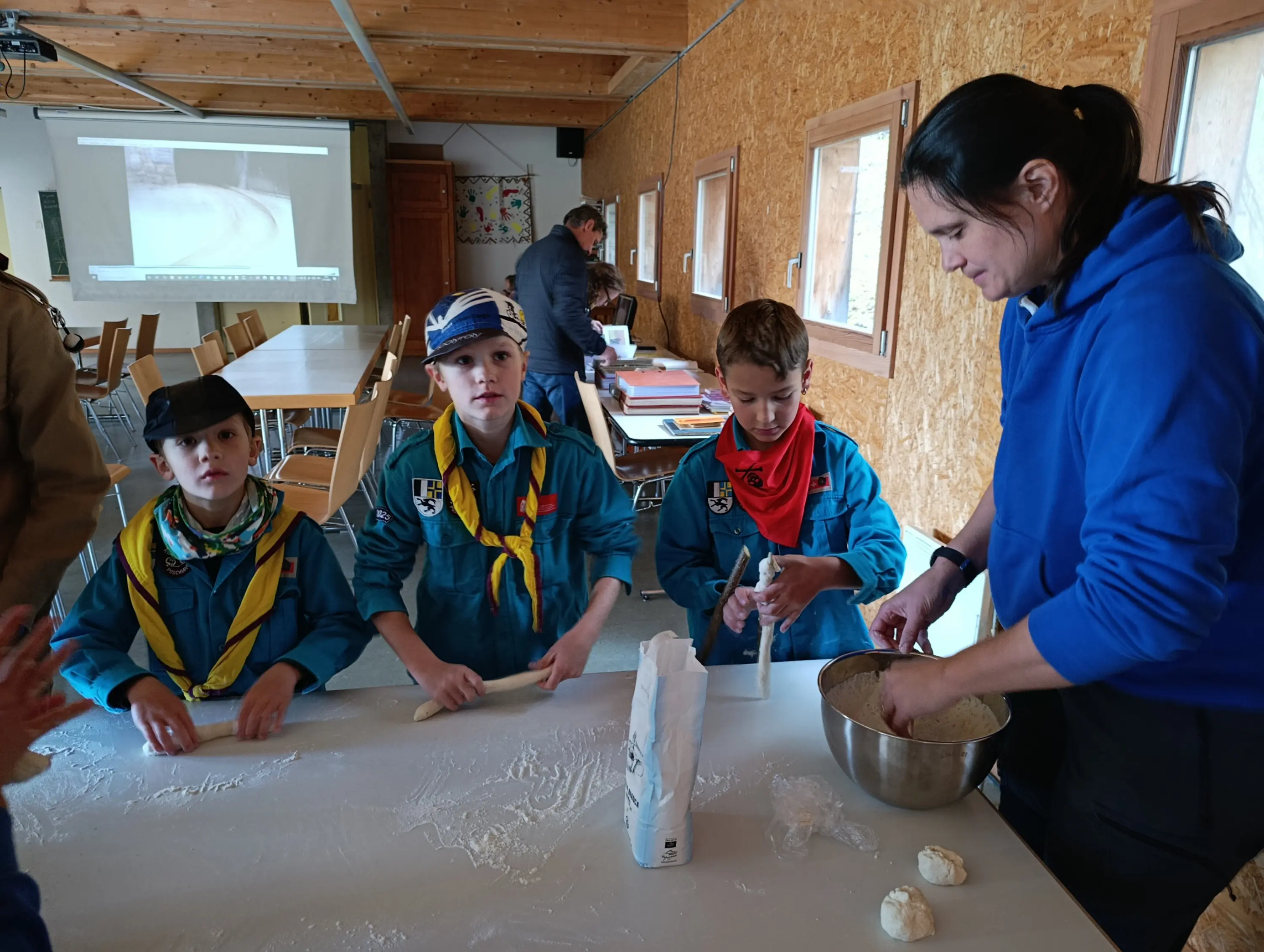 Un gruppo di bambini scout in uniforme blu e sciarpe colorate sta preparando la pasta in una sala di attività. Due ragazzi lavorano la pasta mentre una donna li assiste. Sullo sfondo si possono vedere altre persone e un proiettore acceso.