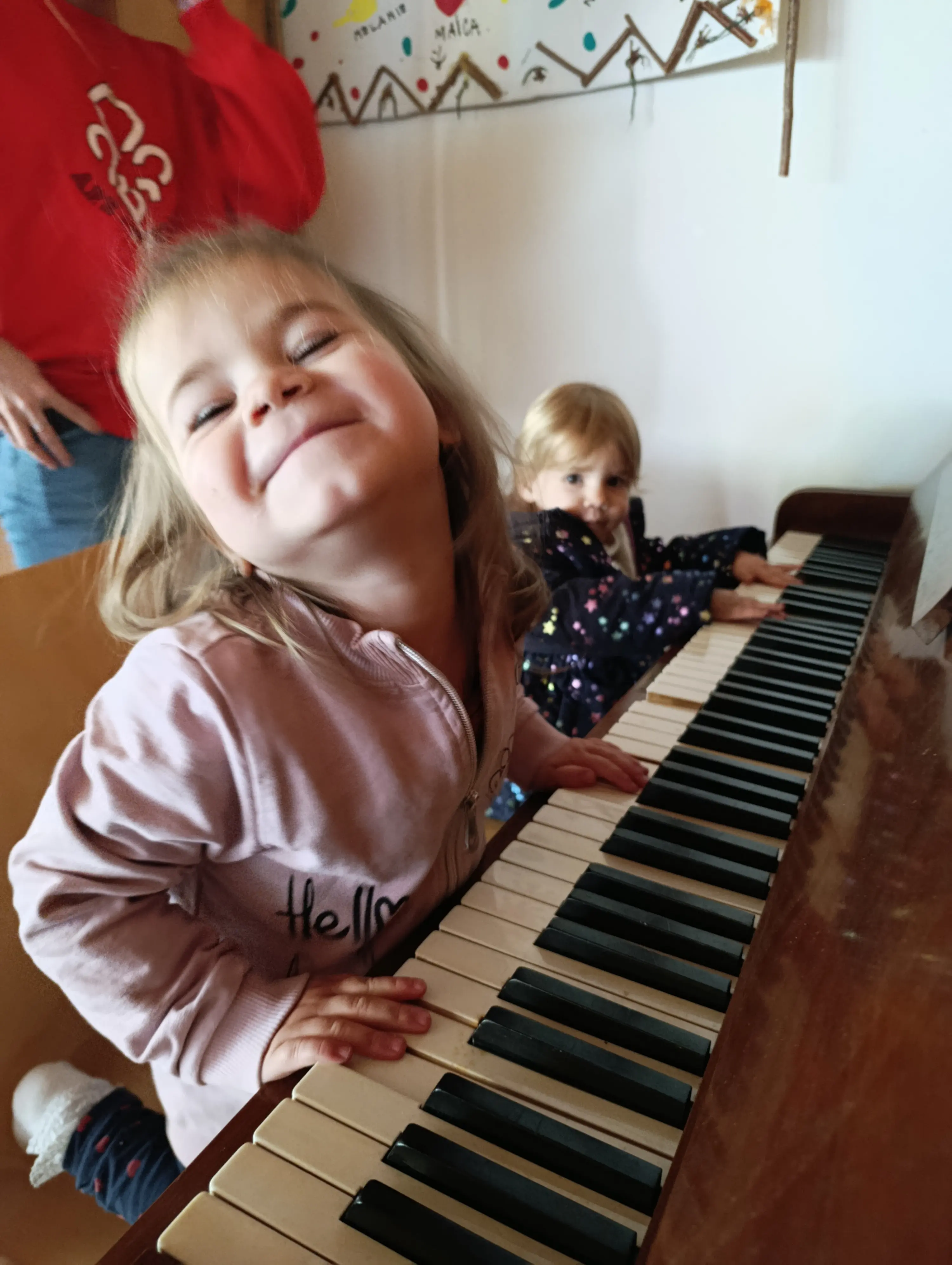 Due bambine sorridono mentre suonano un pianoforte. Una di loro è in primo piano, con un'espressione gioiosa e capelli lunghi. L'altra è sullo sfondo, concentrata sulla musica.