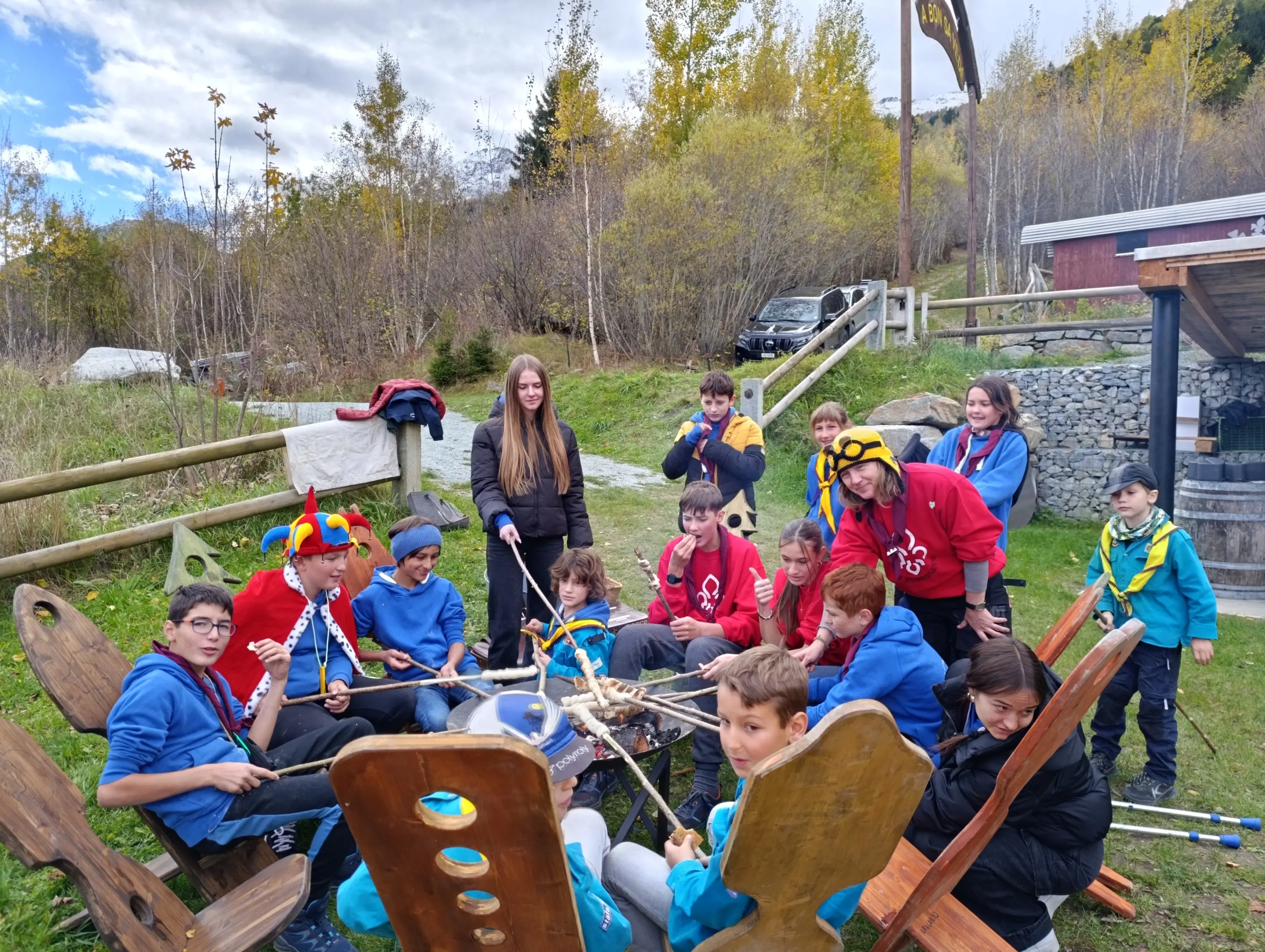 Gruppo di bambini e ragazzi riuniti attorno a un falò all'aperto, sorridenti e impegnati nella preparazione di marshmallow su bastoncini. Alcuni indossano magliette colorate e accessori divertenti. Sullo sfondo, alberi e una struttura in legno.