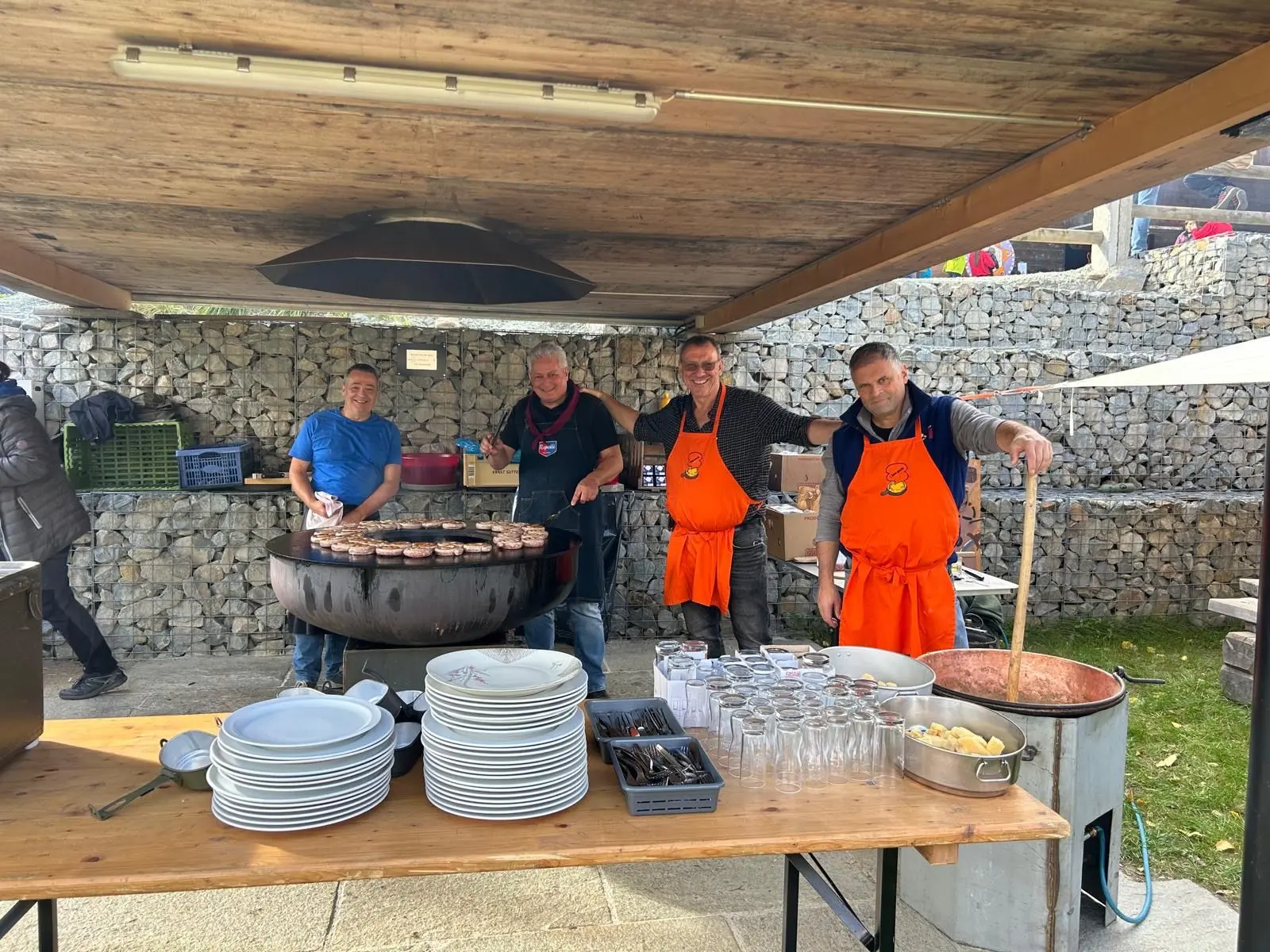 Quattro uomini con grembiuli arancioni cucinano su una griglia sotto un rifugio all'aperto, circondati da piatti e bicchieri. Sul fuoco si vedono hamburger in cottura.