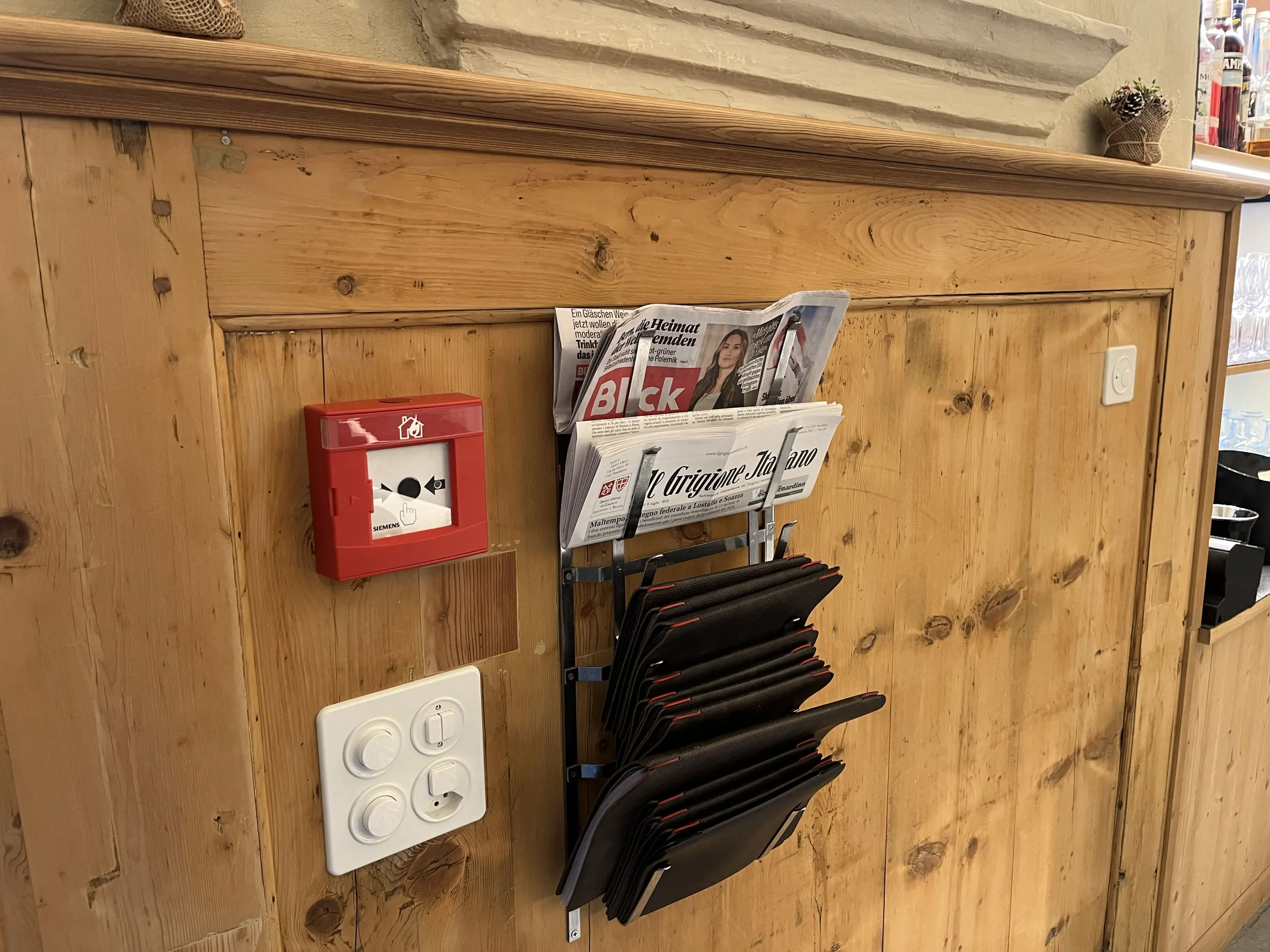 Un muro di legno chiaro con un portagiornali contenente un giornale intitolato "Blick". Sotto ci sono vari menu neri appesi. A sinistra,