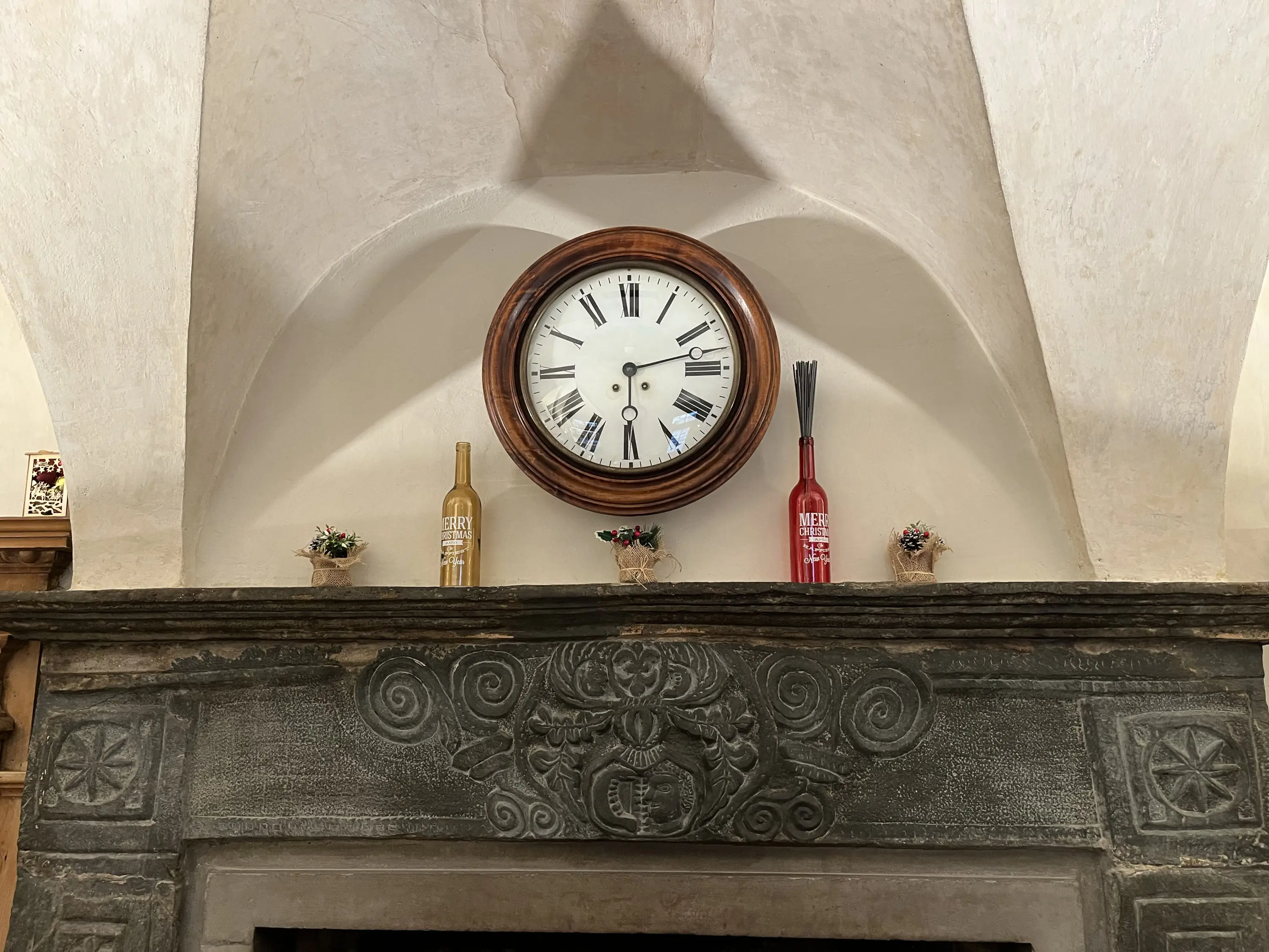 Orologio in legno rotondo con numeri romani appeso sopra un camino in pietra, adornato con tre bottiglie colorate e piccoli fiori in vaso.