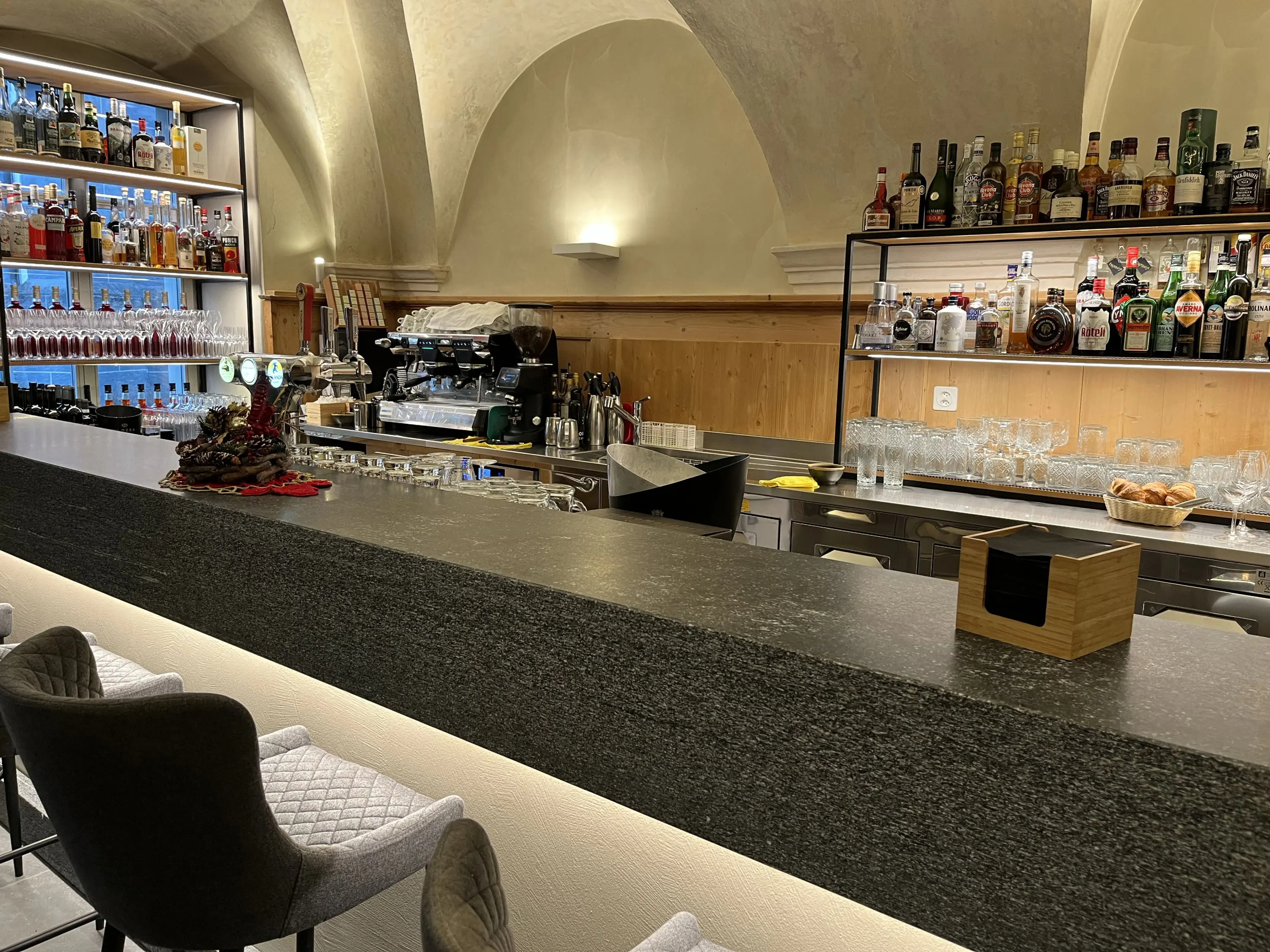 Bar elegante con un lungo bancone in pietra, scaffali retroilluminati pieni di bottiglie di alcolici e bicchieri. Sulle sedie ci sono cuscini gr