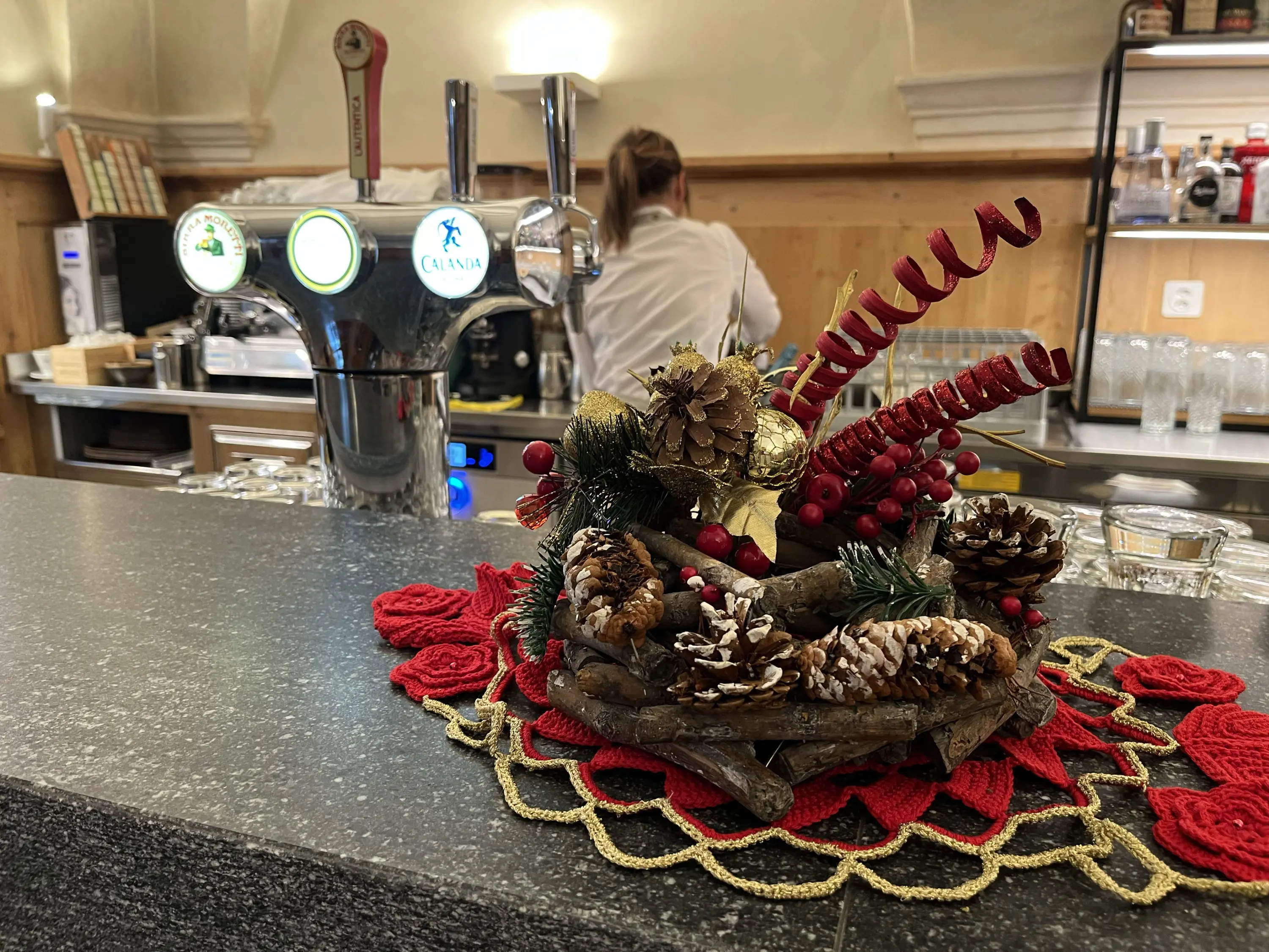 Decorazioni natalizie con pigne e rametti rossi su un tavolo. Sullo sfondo, un bancone bar con una macchina per la birra.