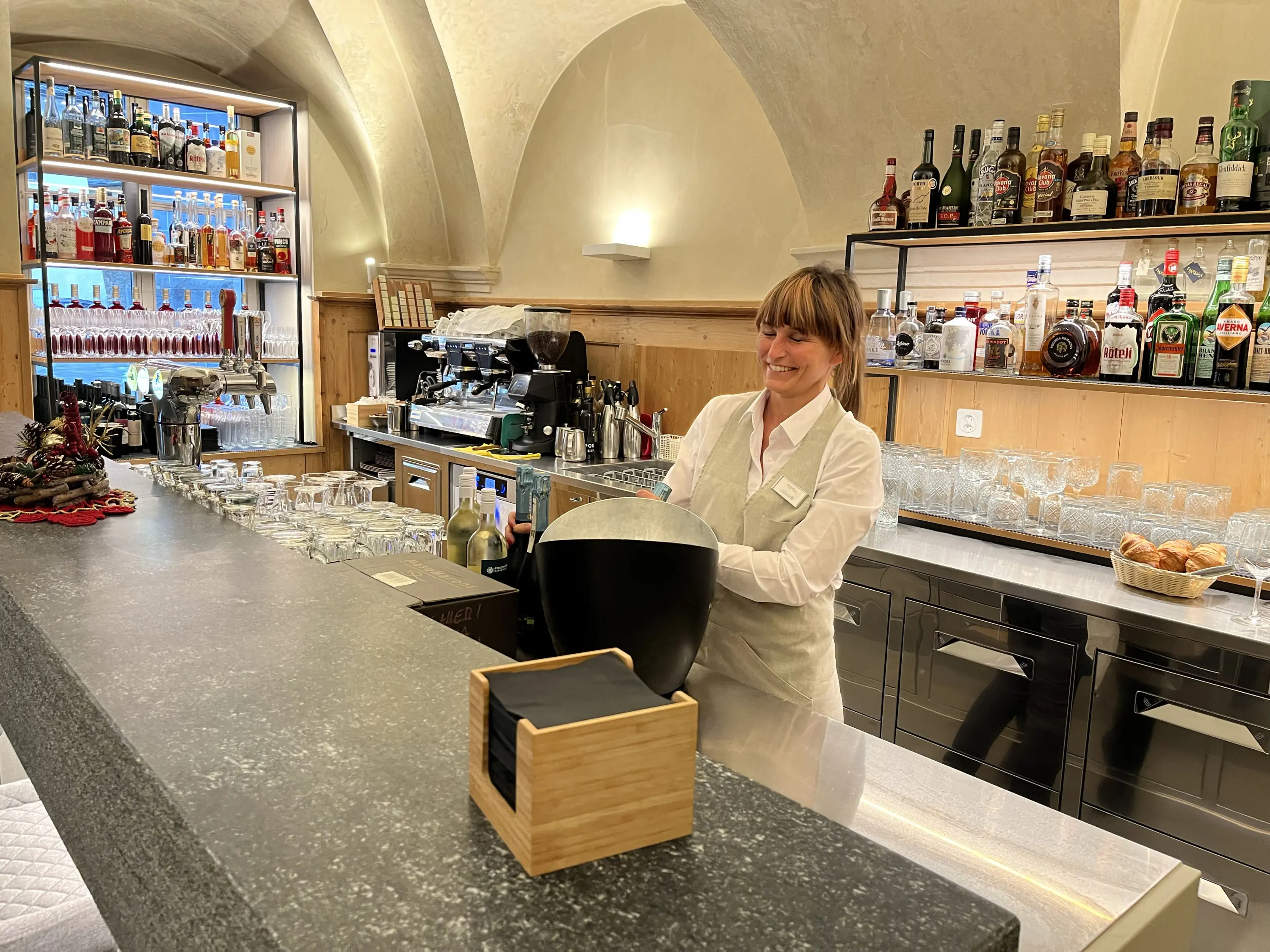 Una bartender sorride mentre lavora al bancone di un bar elegante, con scaffali pieni di bottiglie sullo sfondo e un ambiente accogliente.