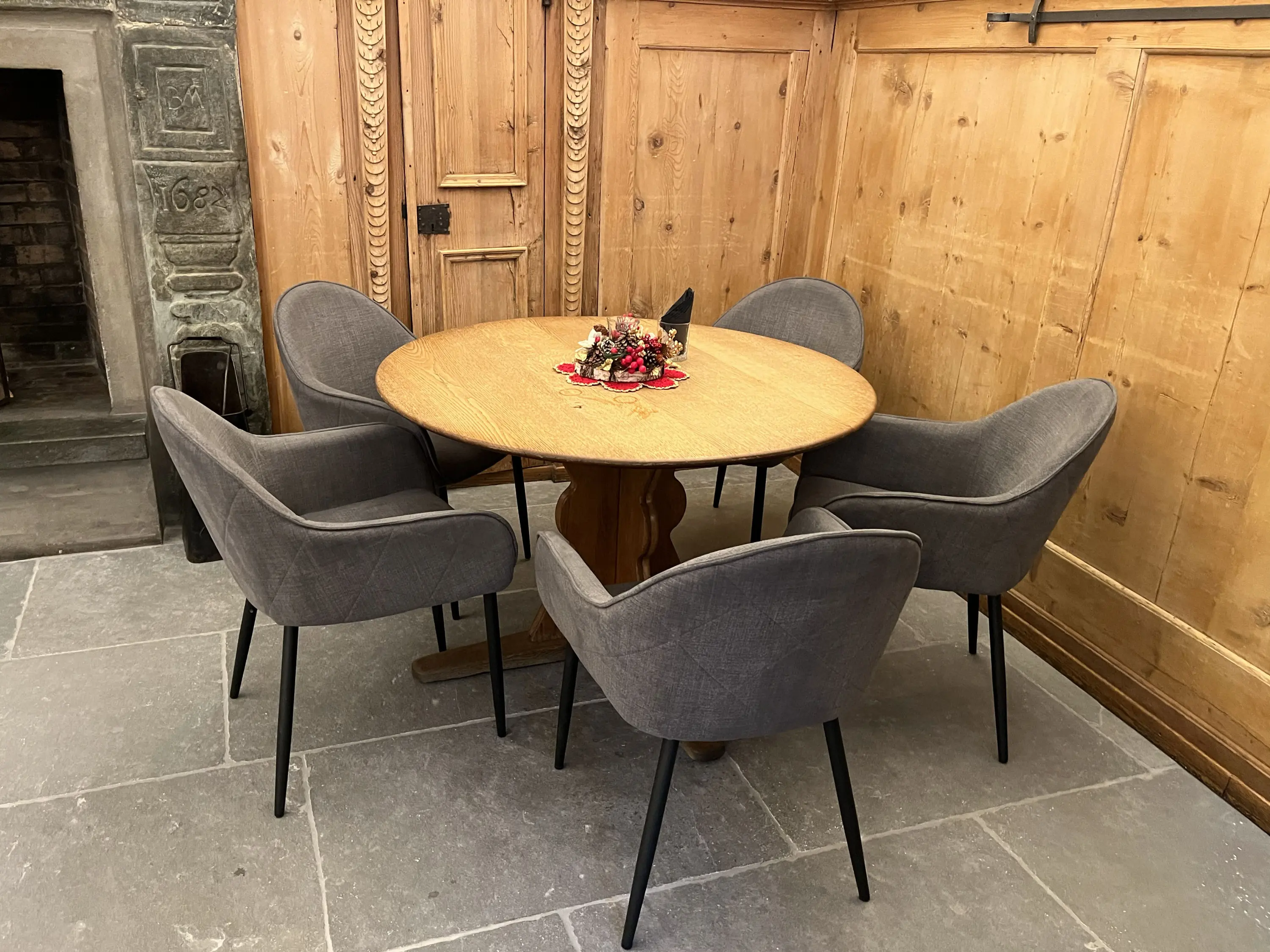 Tavolo rotondo di legno con quattro sedie grigie, situato in un ambiente con pareti in legno e pavimento in pietra. Al centro del tavolo