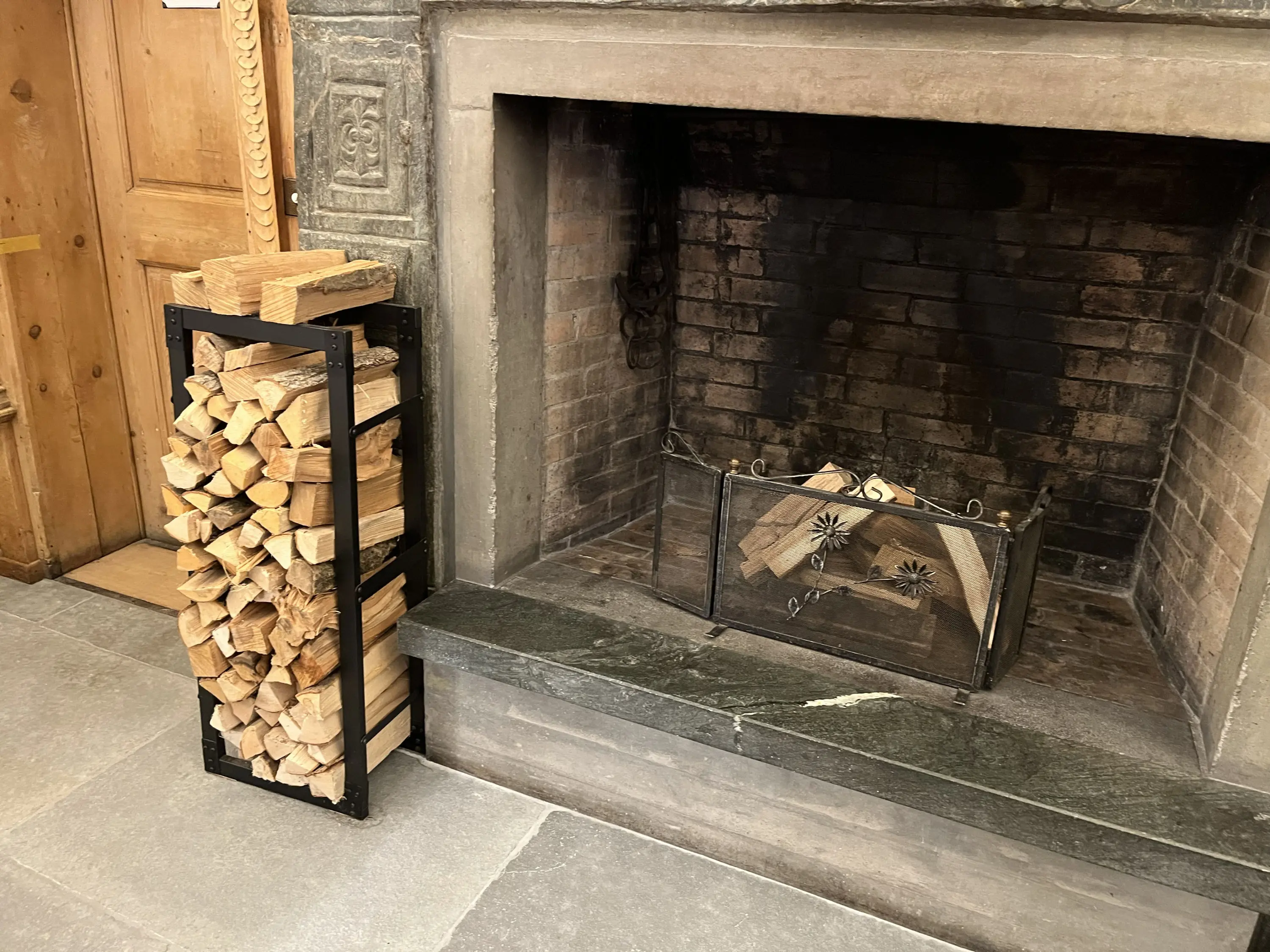 Un supporto di legno contiene legna accatastata accanto a un camino vuoto con una grata di protezione.