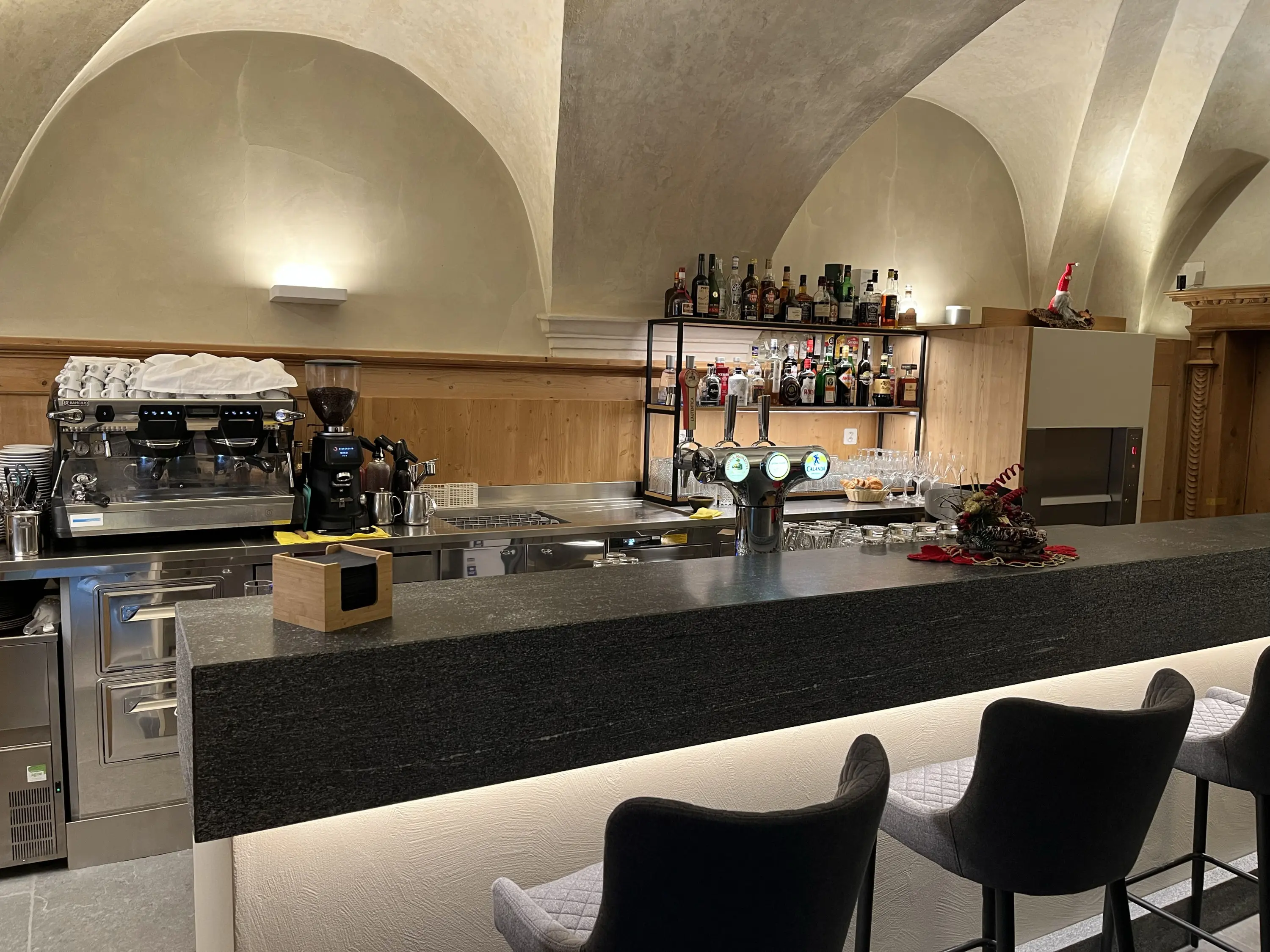 Bar elegante con un bancone in pietra scura, macchine da caffè e una selezione di alcolici. Dietro il bar, scaffali con bottiglie di liquori