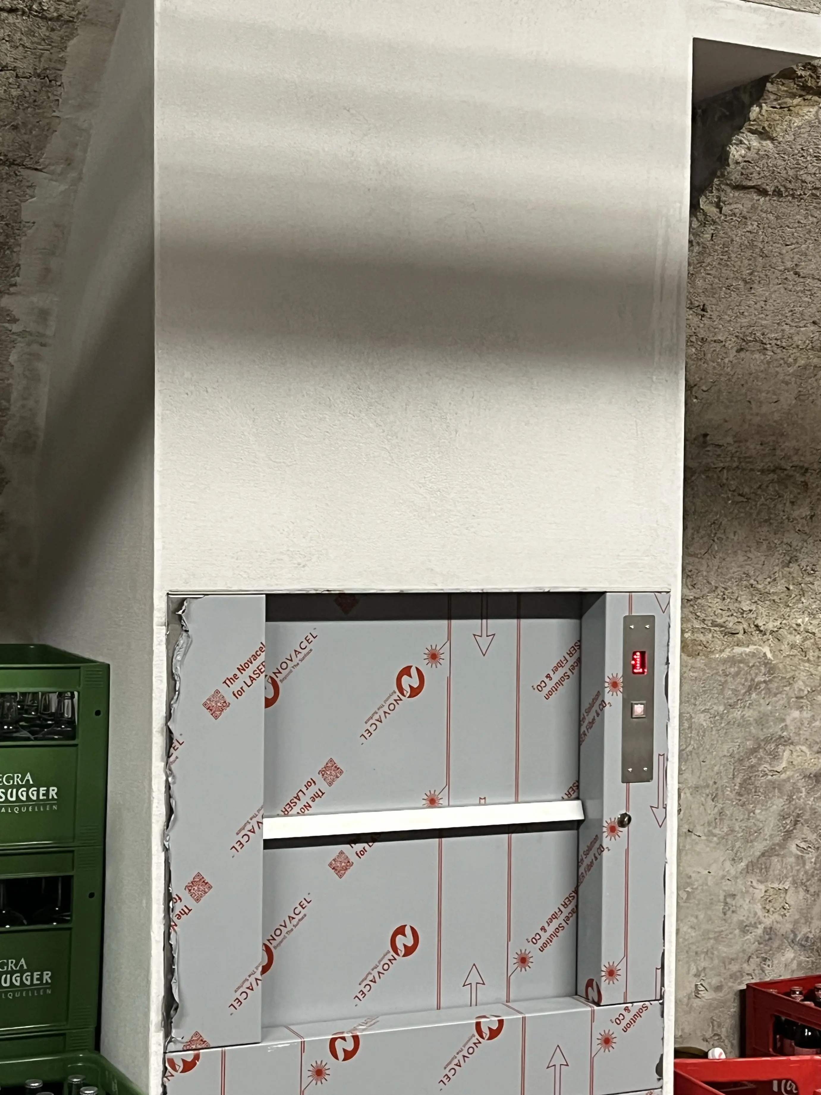 Un ascensore per merci installato in una parete, con la porta chiusa e protetta da un rivestimento in cartone. La parete su cui è montato è fin