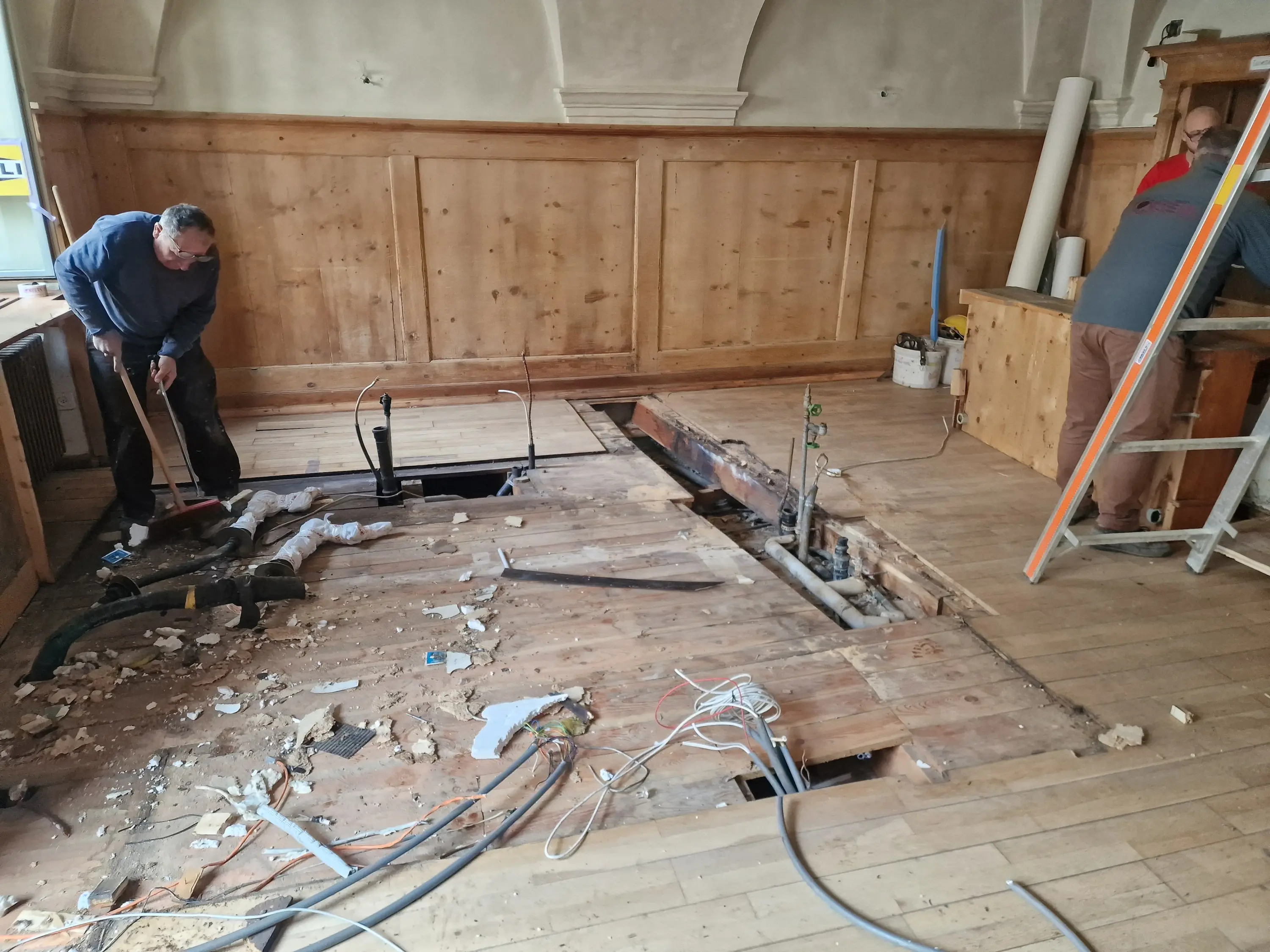 Due operai stanno lavorando in un ambiente con pavimento in legno. Una porzione del pavimento è stata rimossa, mostrando tubi e cavi sottostanti. Attrezzi