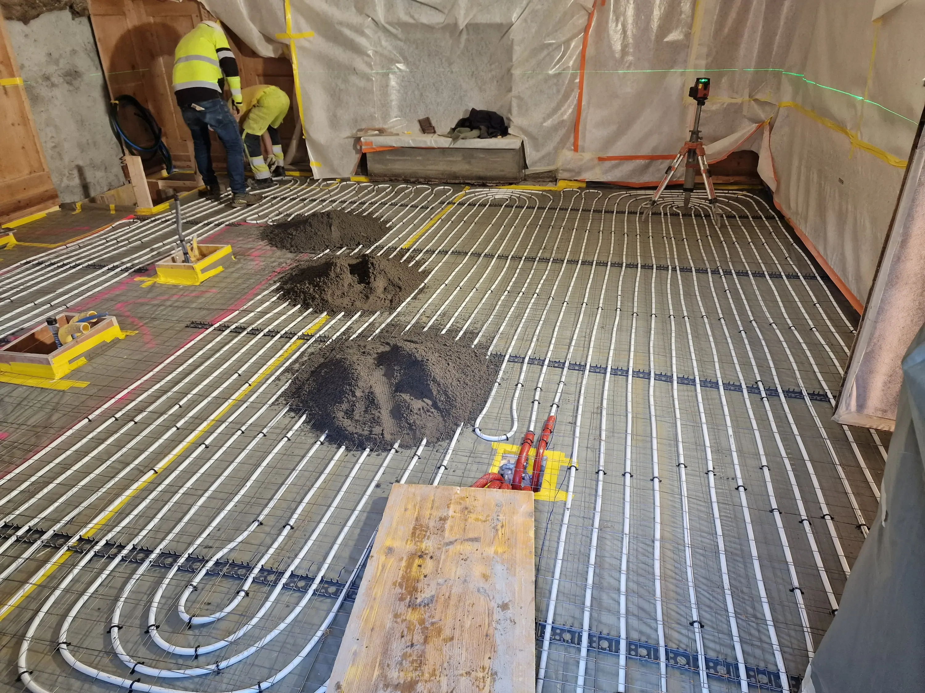 Pavimento di un cantiere con tubi bianchi per riscaldamento a pavimento e cumuli di terra. Due operai in giubbotti riflettenti lavorano sul