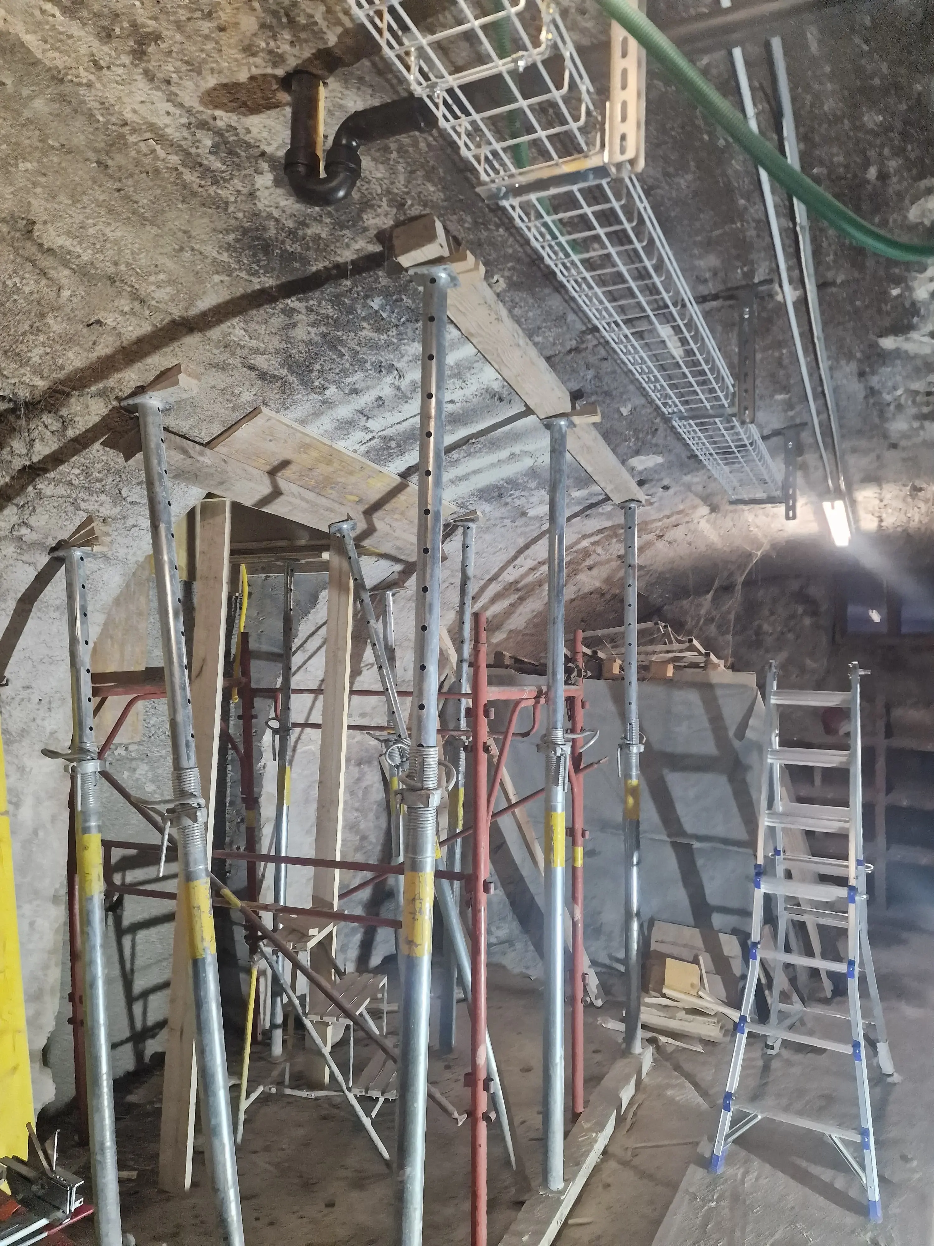 Interno di uno spazio di lavoro sotterraneo con strutture di supporto in metallo, scale e materiali da costruzione. La luce artificiale illumina l'area, evidenziando