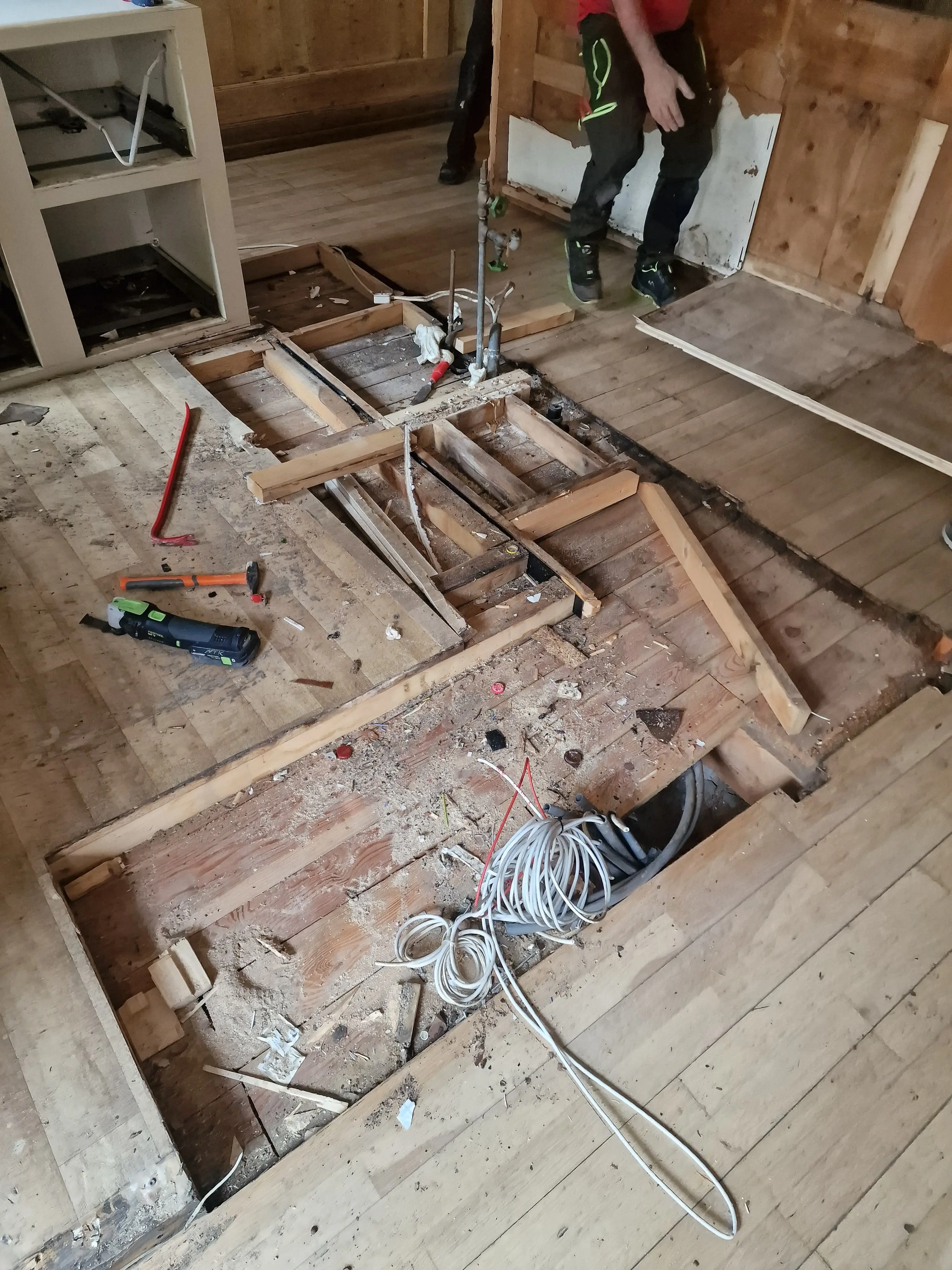 Un pavimento in legno parzialmente smontato, con assi di legno rimosse e visibili cavi e tubature. Attrezzi da lavoro sparsi intorno