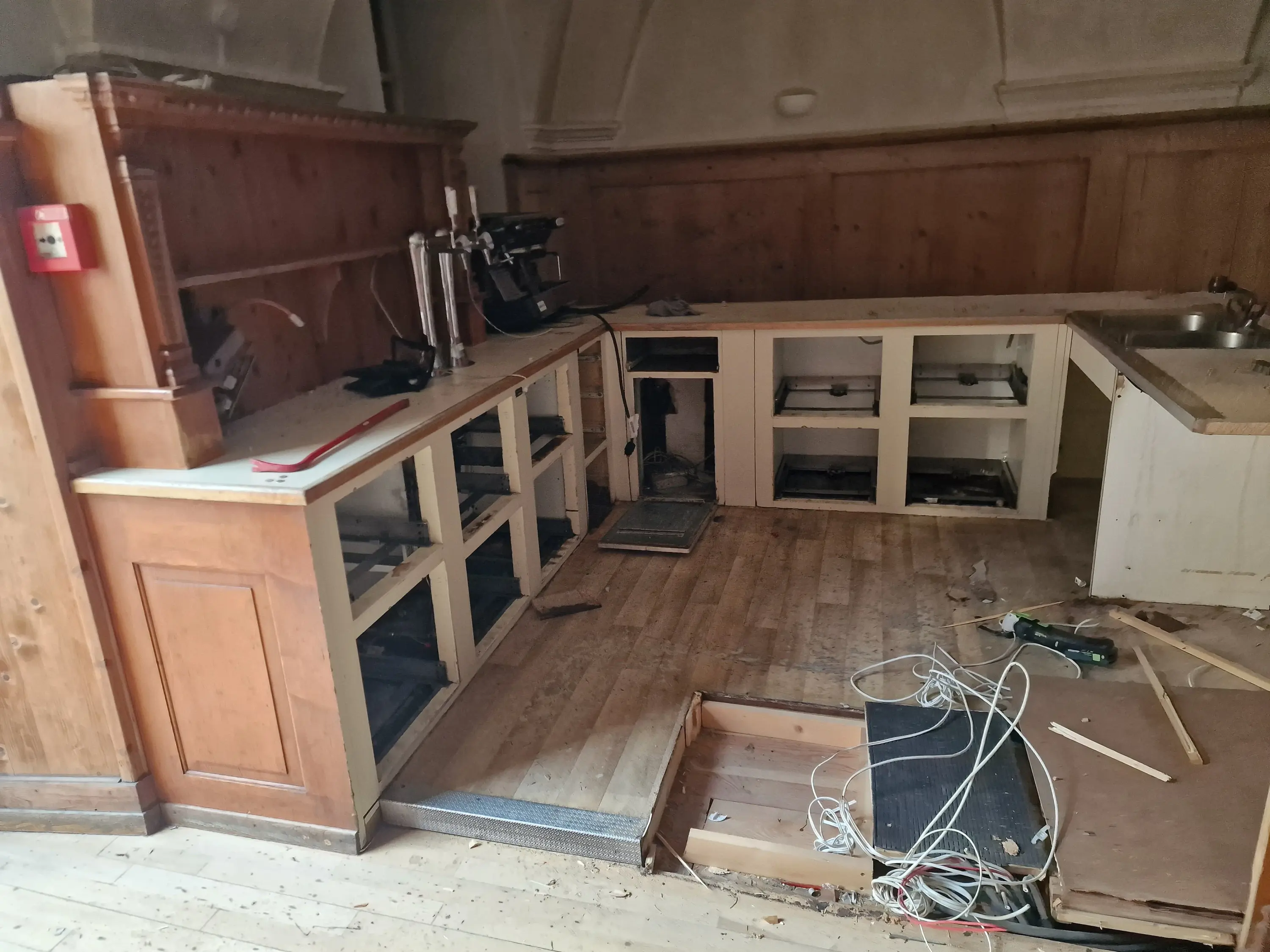 Cucina abbandonata con mobili danneggiati, ripiani vuoti e fili elettrici disordinati. Pavimento in legno visibilmente usurato.