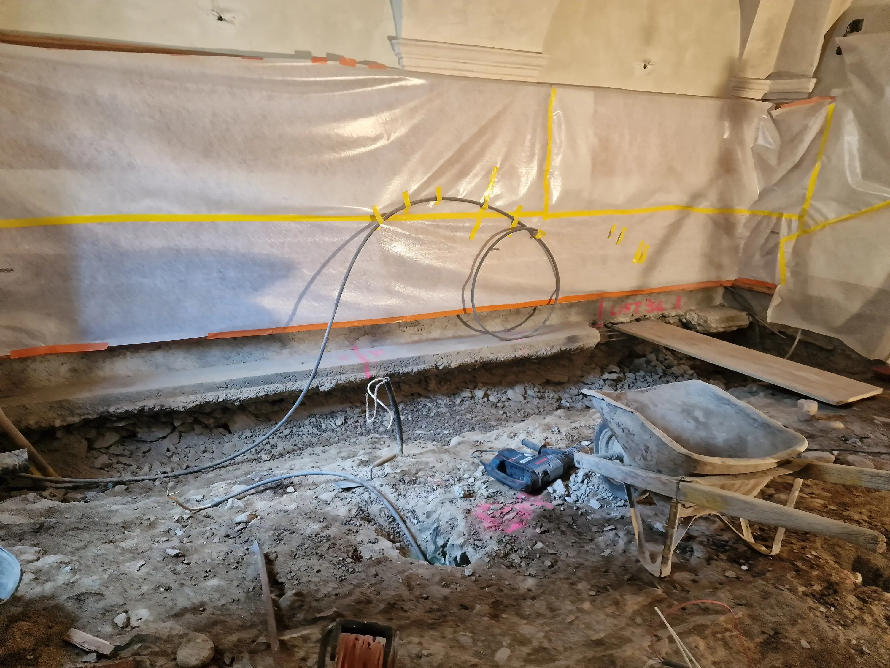 Ambiente di costruzione con pareti protette da teli trasparenti. Un'area con terreno scavato e una carriola presente, mentre cavi elettrici sono visibili sopra