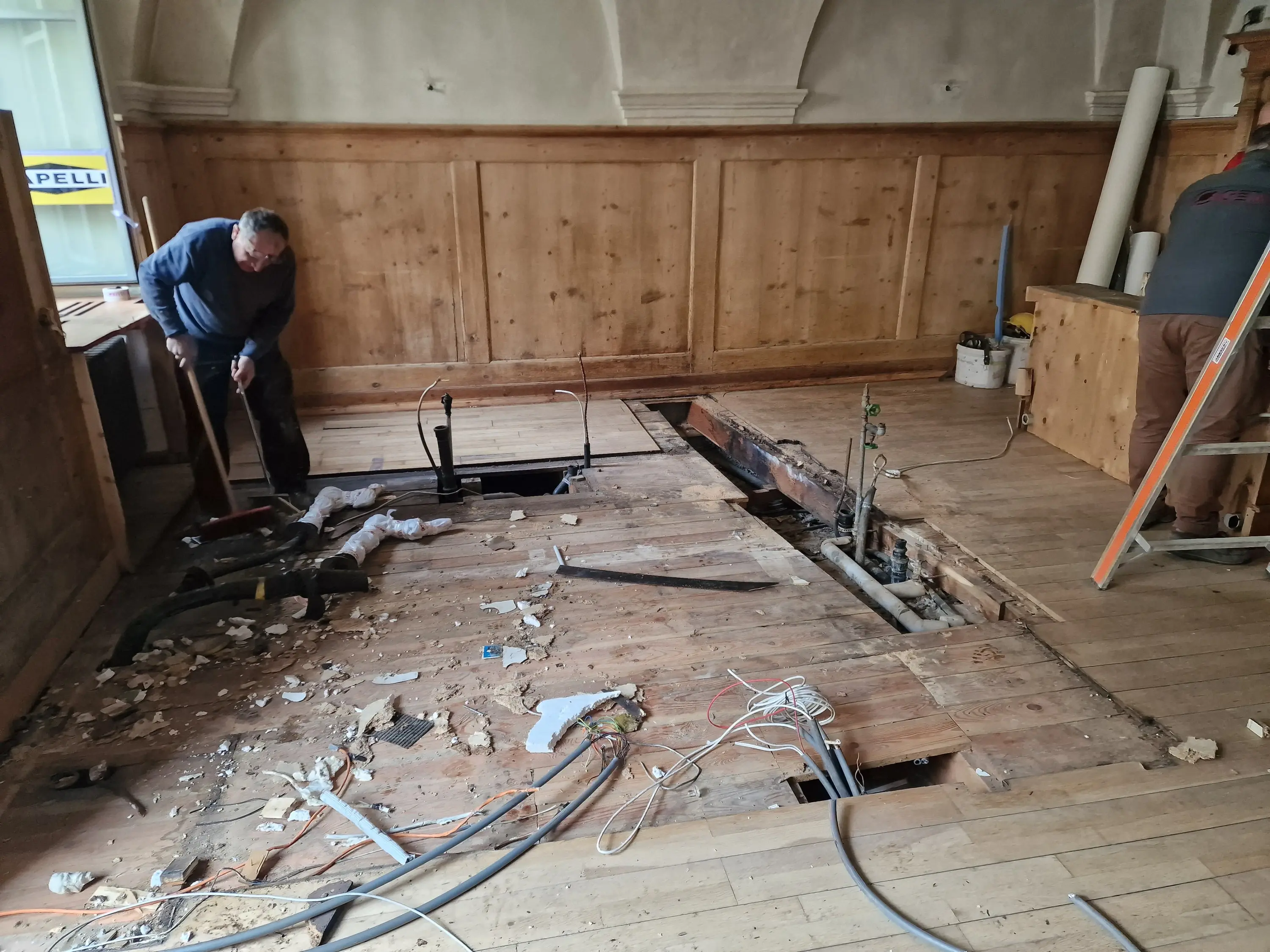 Un uomo sta lavorando sul pavimento di legno, rimuovendo tavole e mostrando un'apertura nel pavimento. Due tubi e cavi elettrici sono visibili sotto