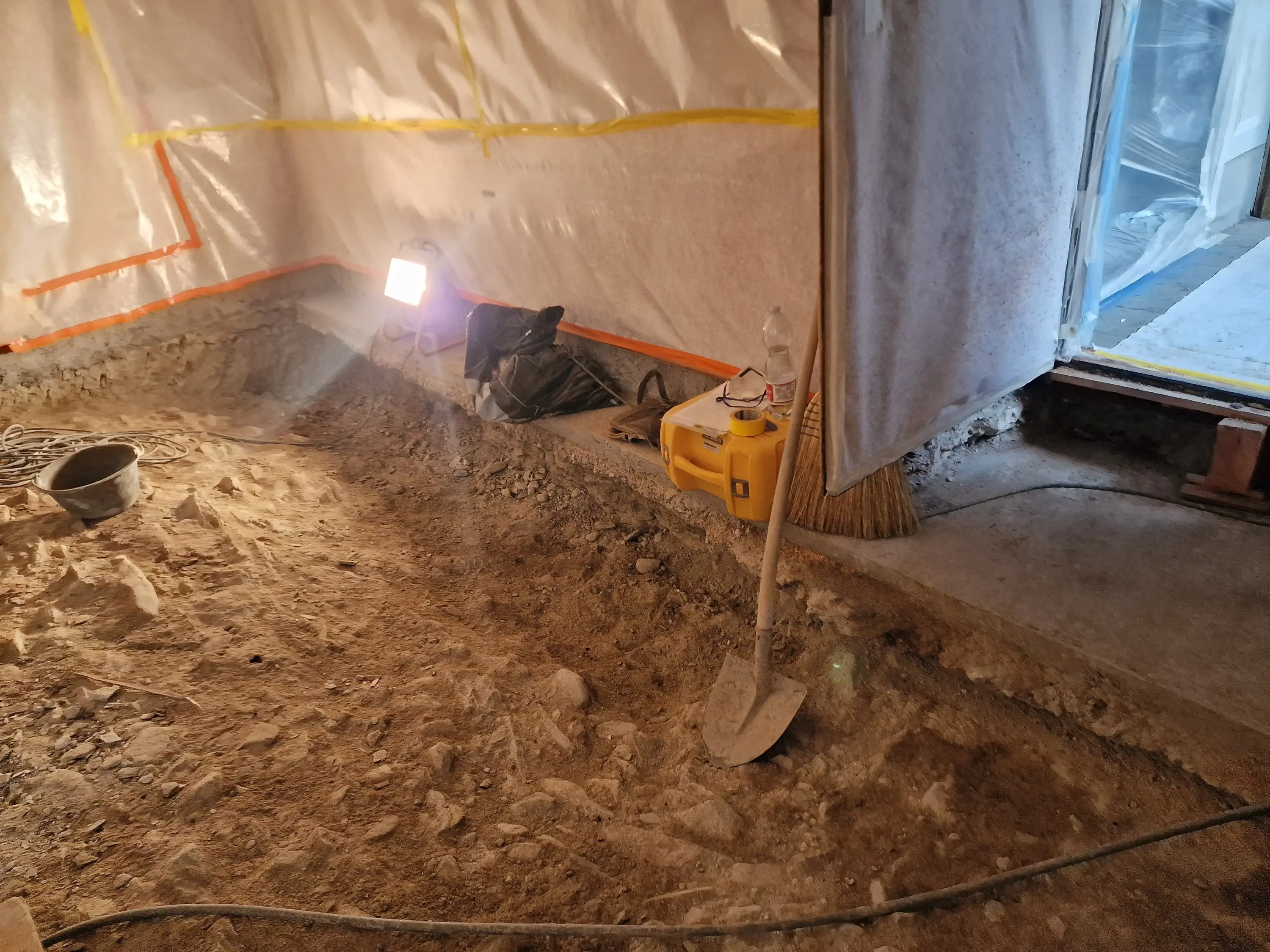 Un cantiere in fase di ristrutturazione con sacchi di materiali, attrezzi e un faretti accesi su un pavimento di terra. Una scopa è appoggiata