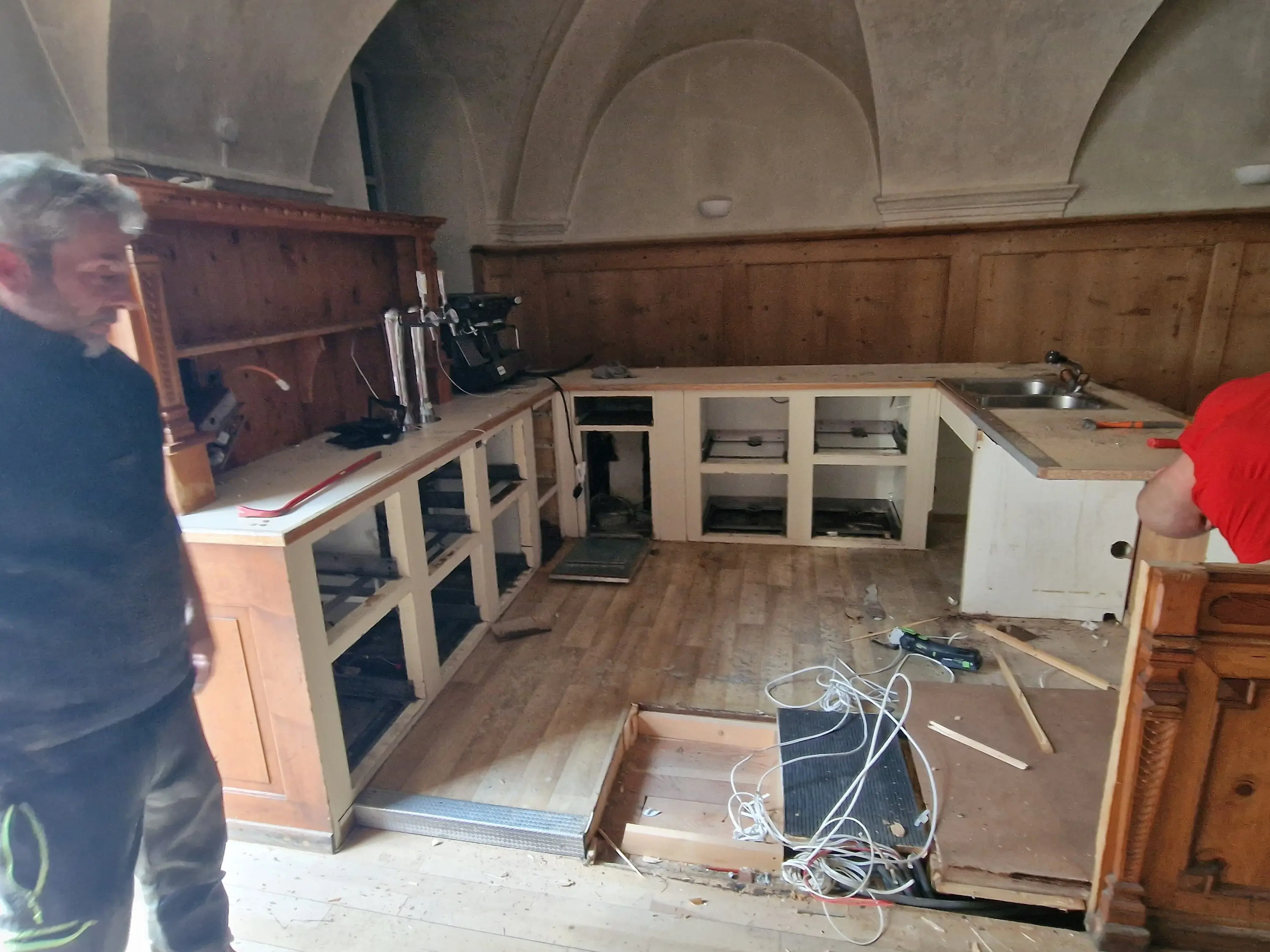 Una cucina in fase di ristrutturazione con armadi incompleti e attrezzature da cucina in disordine. Due uomini stanno lavorando nel locale.