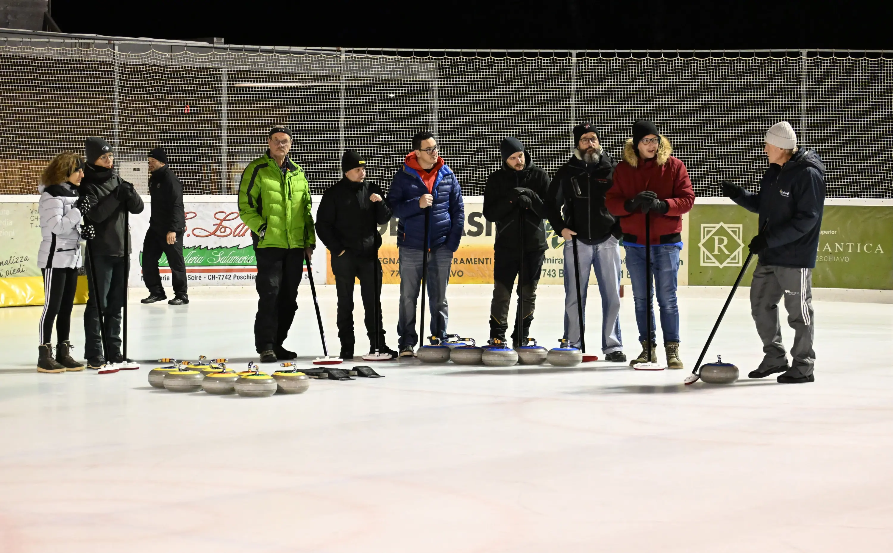 Gruppo di persone in abbigliamento invernale su una pista di curling, alcune in piedi vicino a una serie di pietre da curling, mentre un uomo spiega le tecniche del