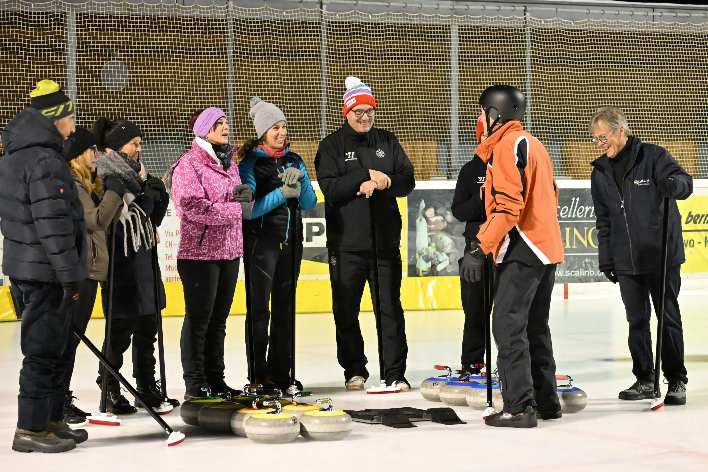 Gruppo di persone in abbigliamento invernale sulla pista di curling, mentre un istruttore spiega le tecniche di gioco. Alcuni partecipanti tengono in mano i bast