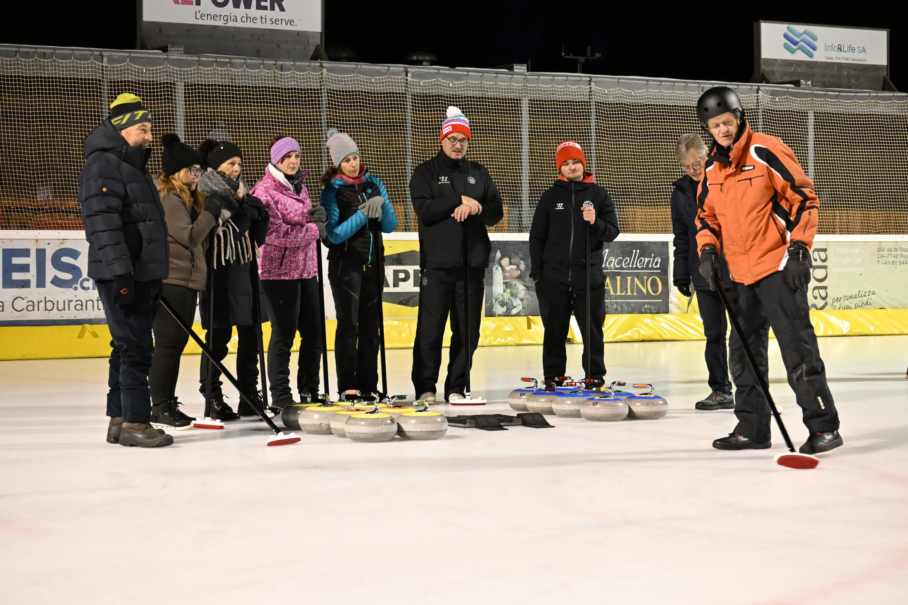 Un gruppo di persone vestite con abbigliamento invernale si trova su un campo da curling. Alcuni partecipanti osservano un istruttore che spiega come giocare, mentre