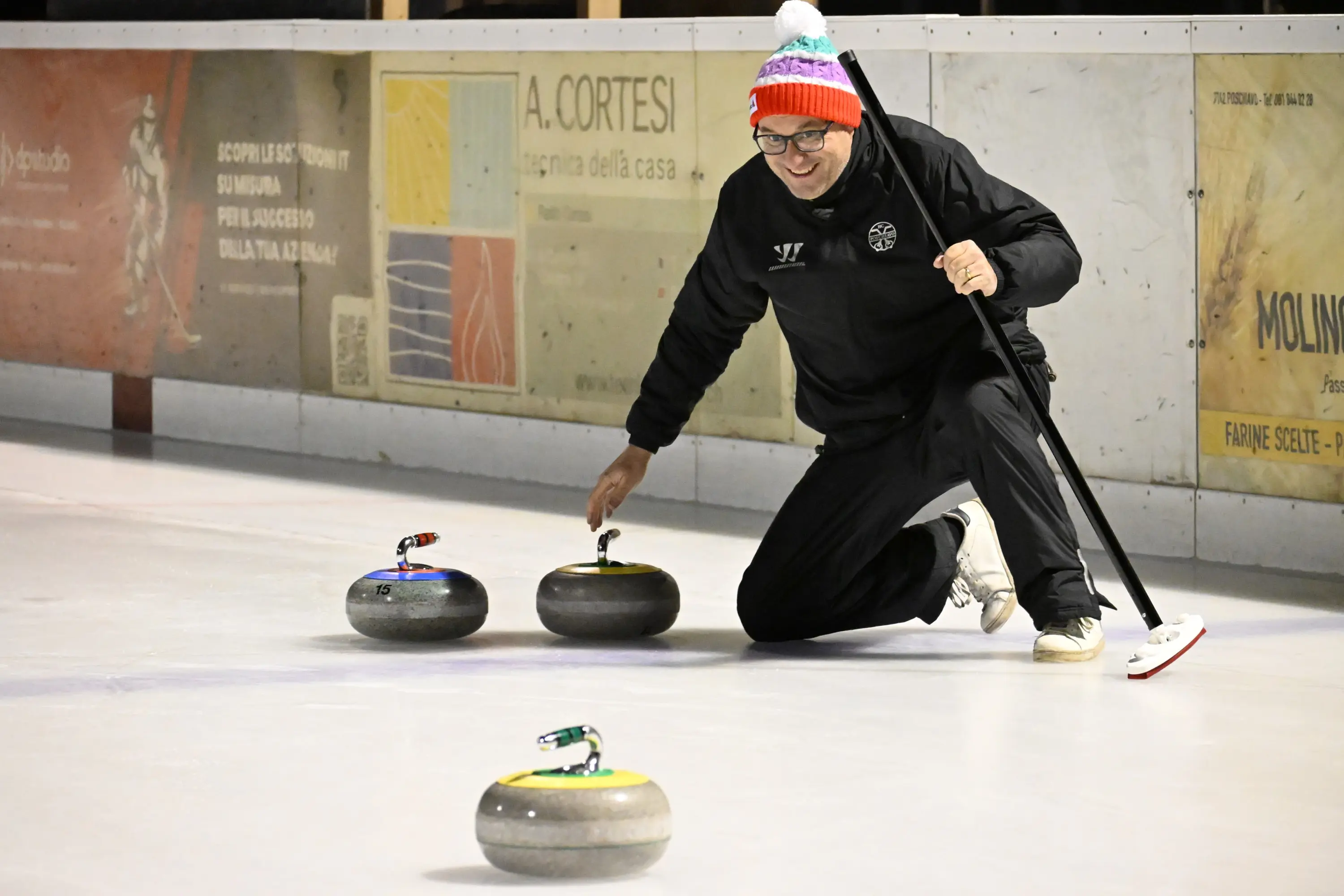 Una persona sorridente in abbigliamento sportivo si inginocchia su una pista di curling, mentre lancia una pietra e osserva attentamente il suo movimento.