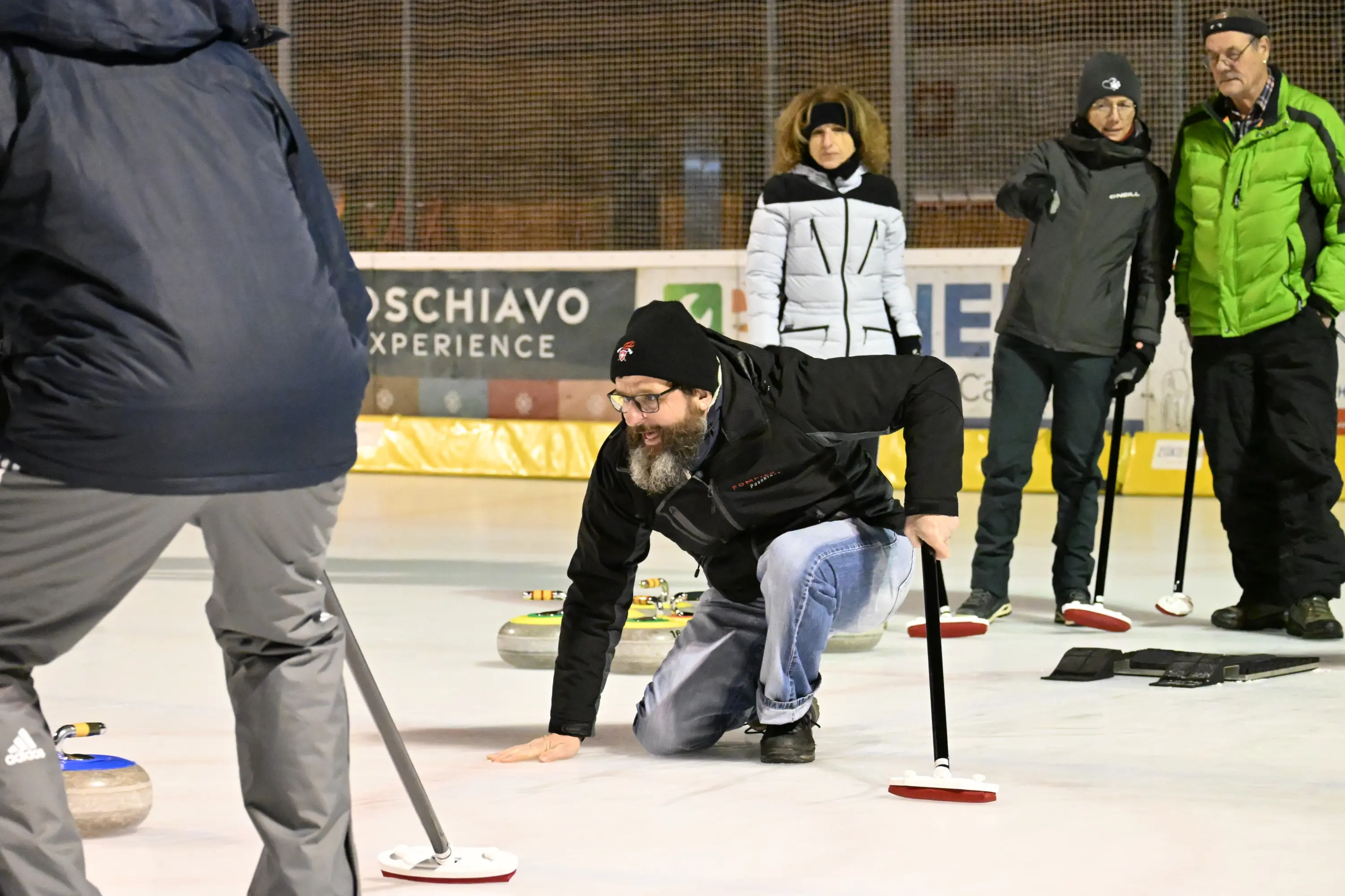 Un uomo con una barba folta si inginocchia su una superficie di ghiaccio mentre lancia una pietra da curling. Altri partecipanti osservano e prendono appunti. La scena