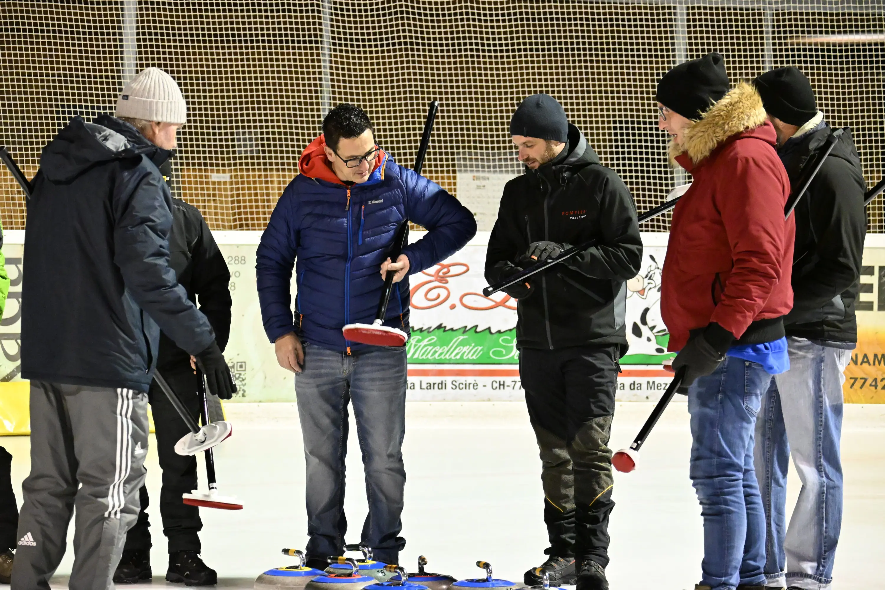 Gruppo di uomini riuniti su un campo di curling, intenti a discutere e manovrare le loro attrezzature. Indossano giacche pesanti e cappelli,