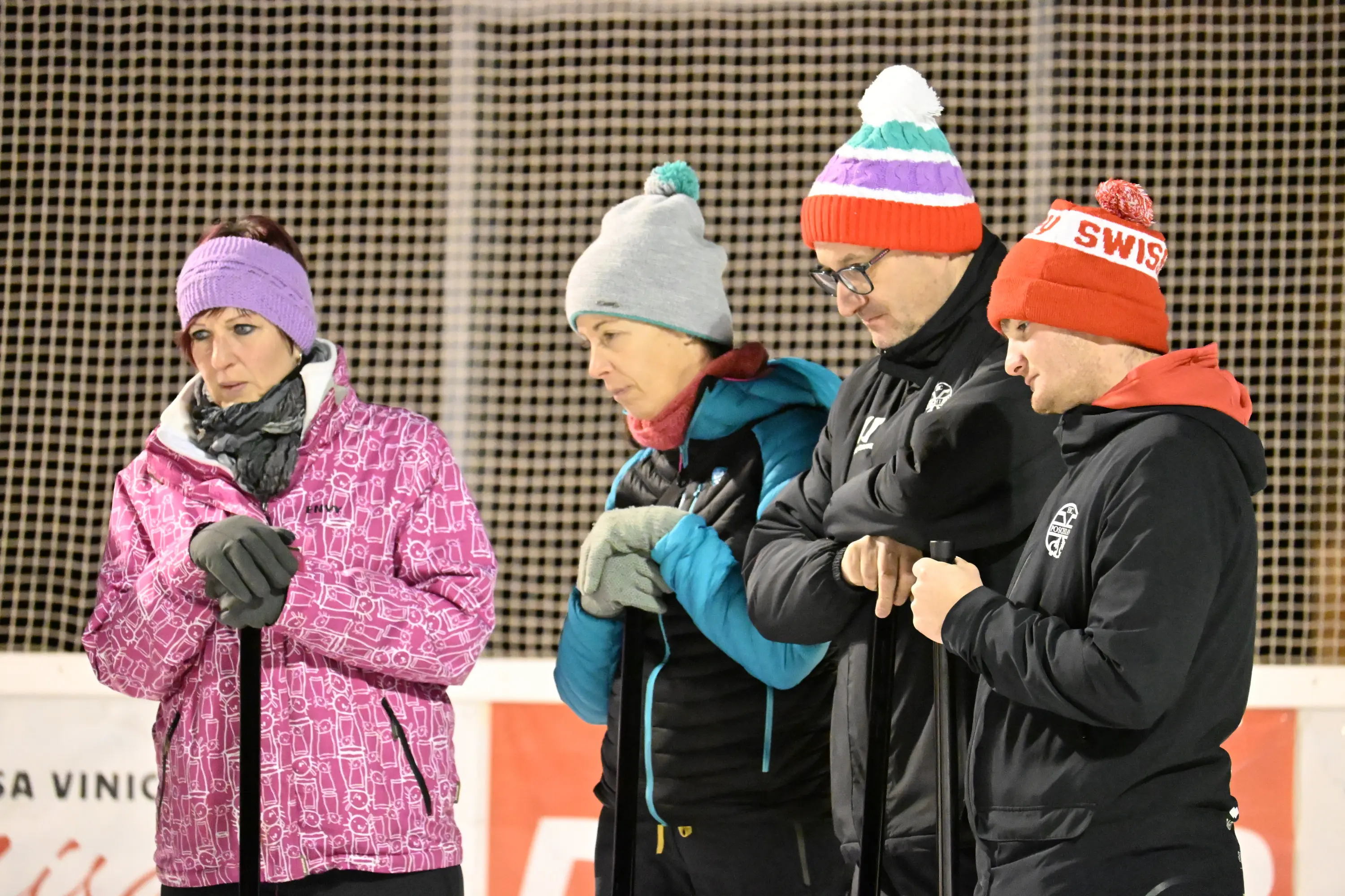 Gruppo di quattro persone in abbigliamento invernale, con cappelli colorati e guanti, che stanno in fila con le loro stecche da curling, guardando verso il