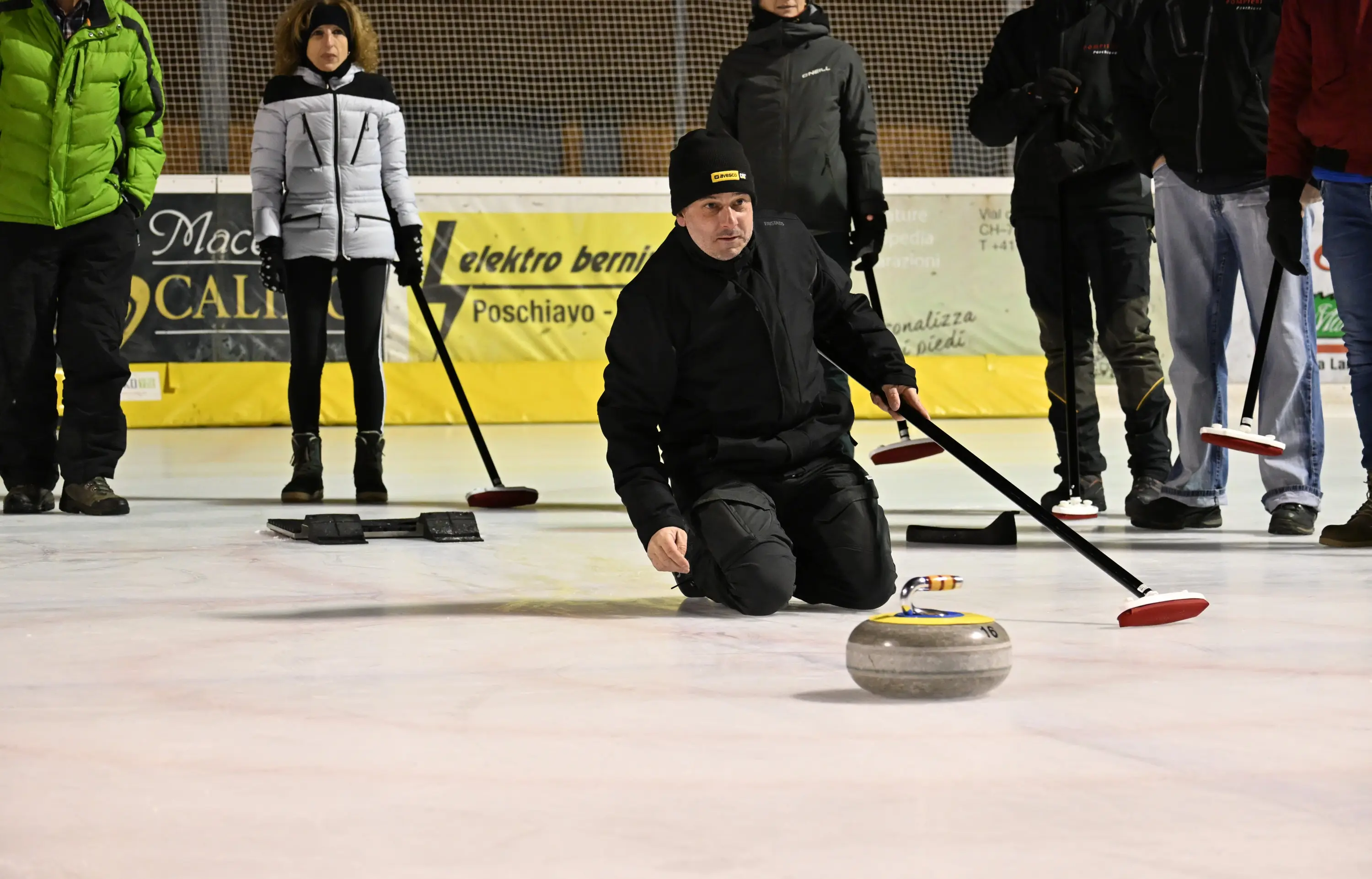 Un uomo in abbigliamento nero si inginocchia sul ghiaccio mentre lancia una pietra da curling. Altre persone osservano in background, alcune sono vestite in giacche