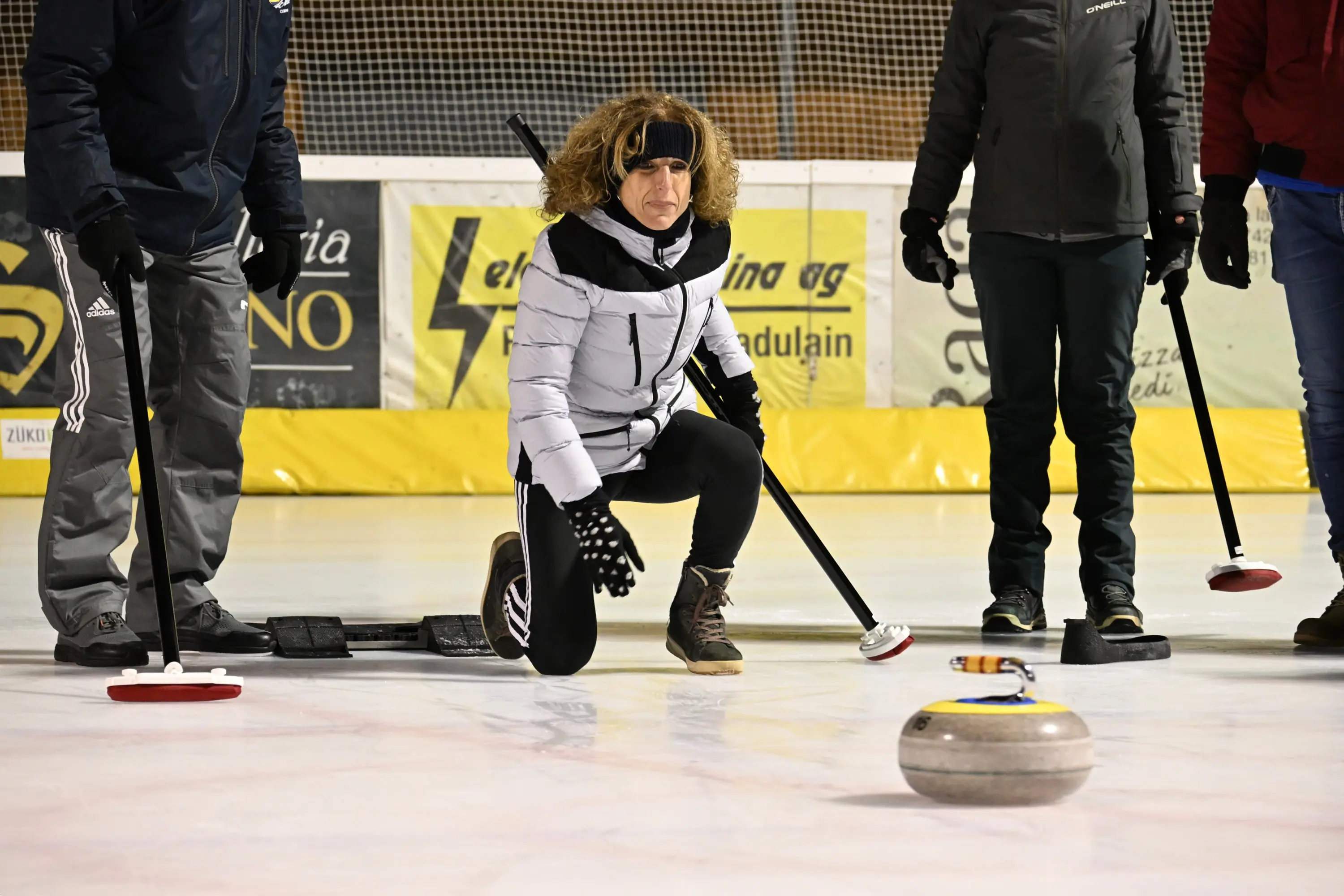 Una donna in abbigliamento invernale si inginocchia sulla pista di curling, mentre osserva attentamente un disco di pietra che scivola sul ghiaccio. Altri partecip
