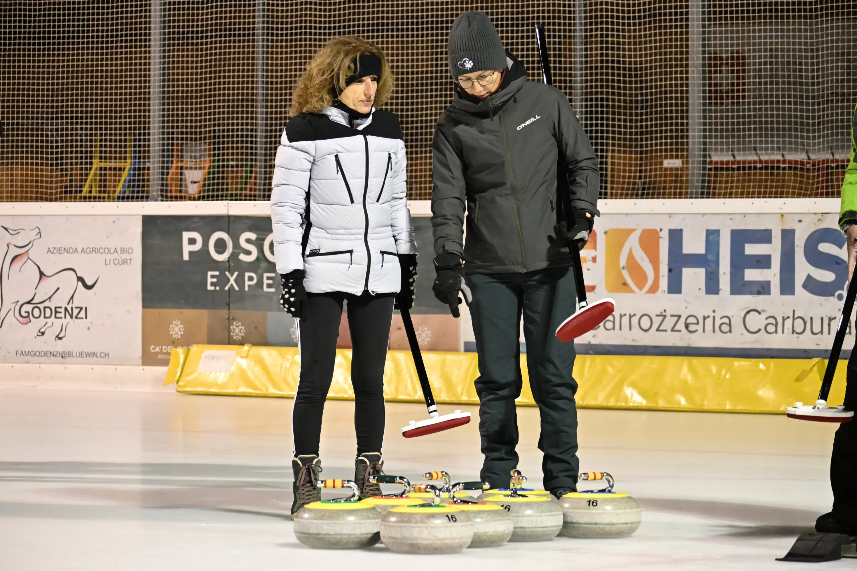Due persone sono sul ghiaccio mentre osservano attentamente delle pietre da curling. Una donna indossa un giubbotto bianco e l'altra persona, un uomo, è vestito con