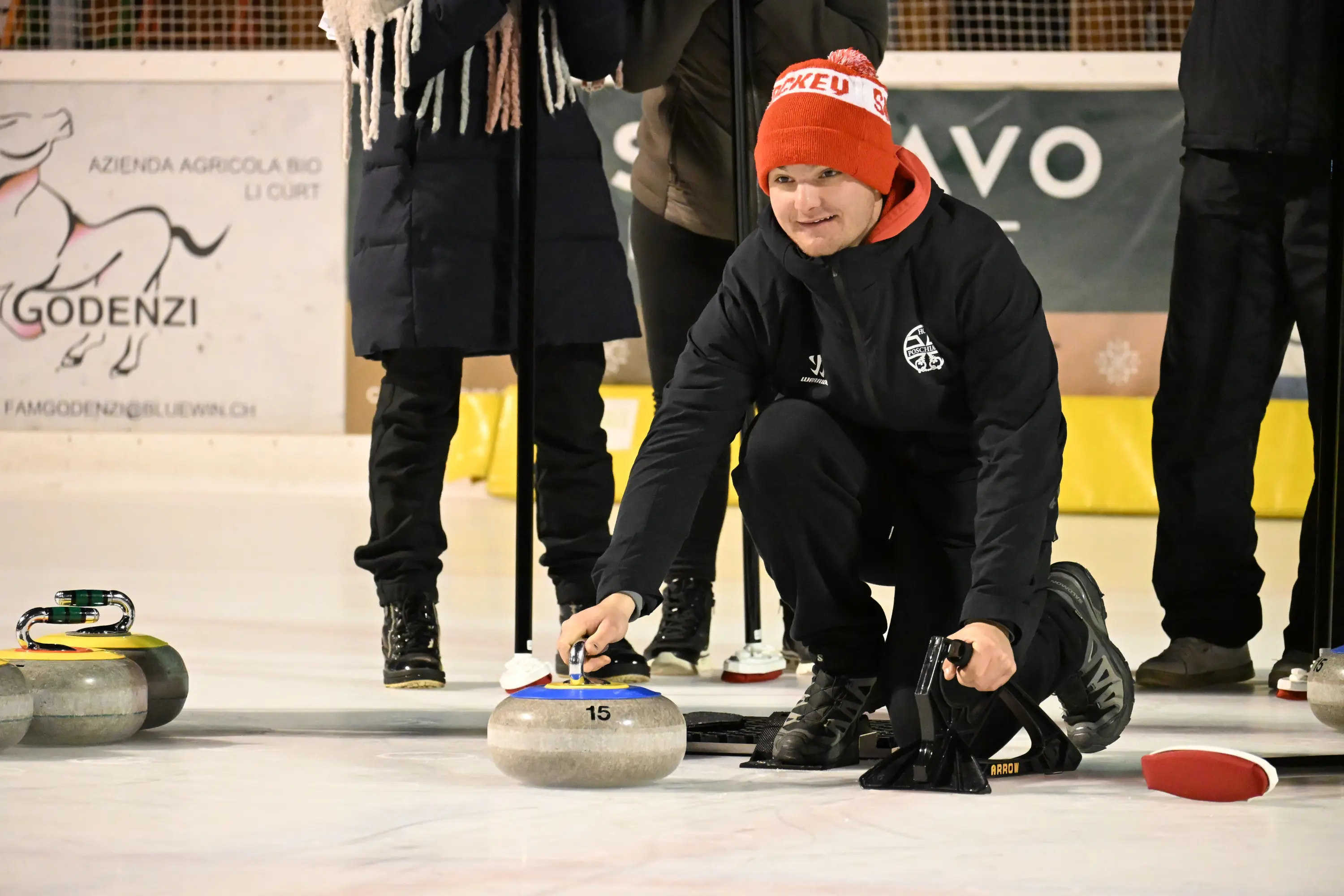 Giocatore di curling in ginocchio mentre si prepara a lanciare una stone sul ghiaccio. Indossa un cappello rosso e un abbigliamento sportivo scuro.