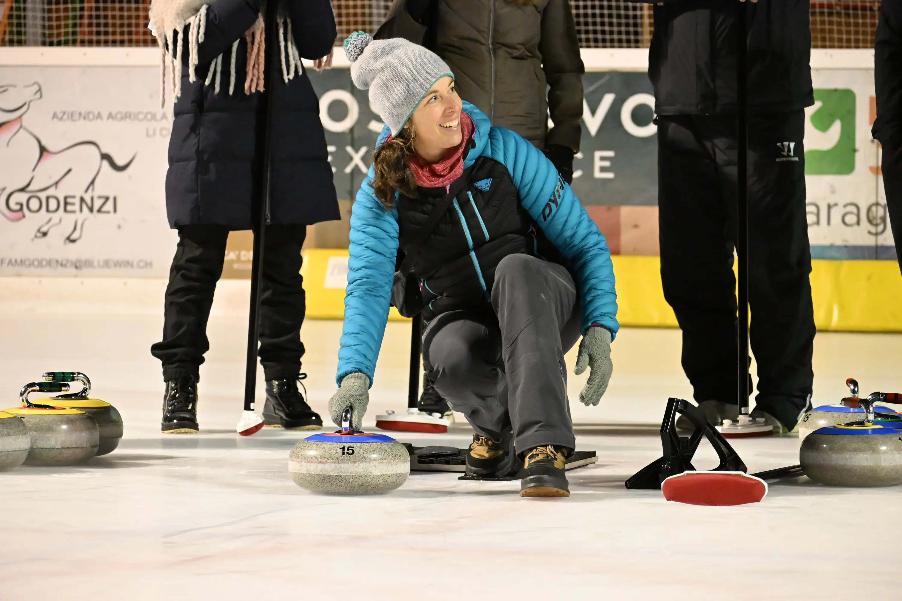 Una donna sorridente indossa un cappello grigio e una giacca blu mentre si abbassa per lanciare una pietra da curling su una pista di ghiaccio. Attorno a