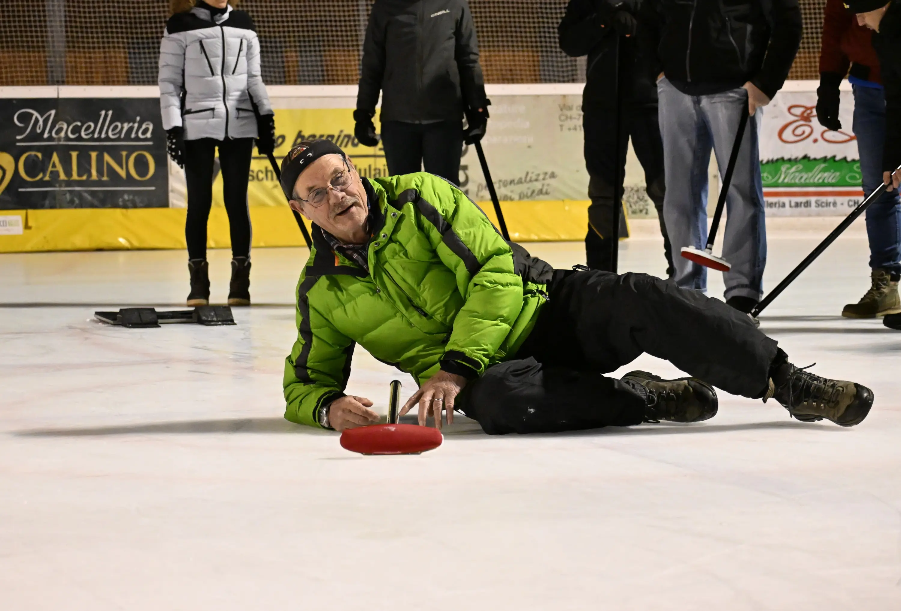 Un uomo anziano indossa una giacca verde mentre si trova sdraiato sul ghiaccio, intento a tirare un disco da curling. Alcune persone lo osservano nel contesto di un