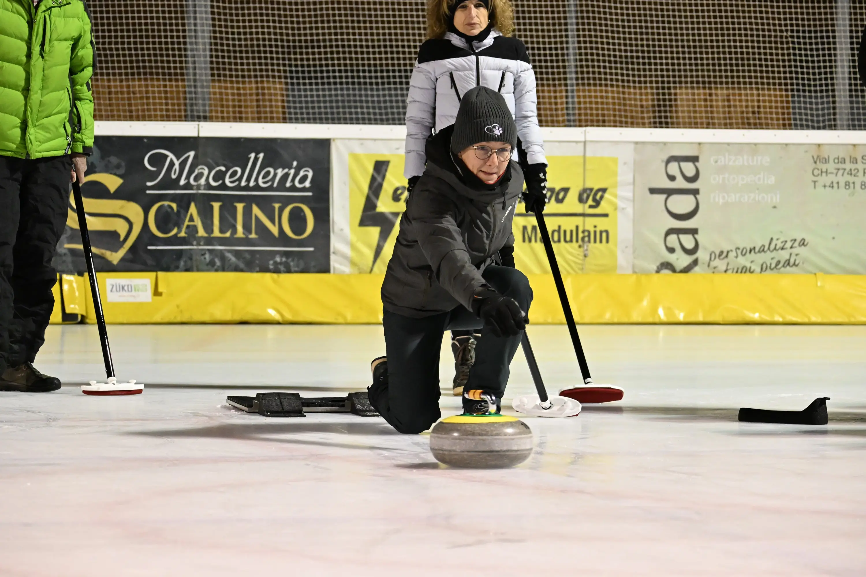 Una persona si concentra mentre lancia una pietra da curling su un campo di ghiaccio. Sullo sfondo, un'altra persona osserva. Gli individui indossano abbigliamento