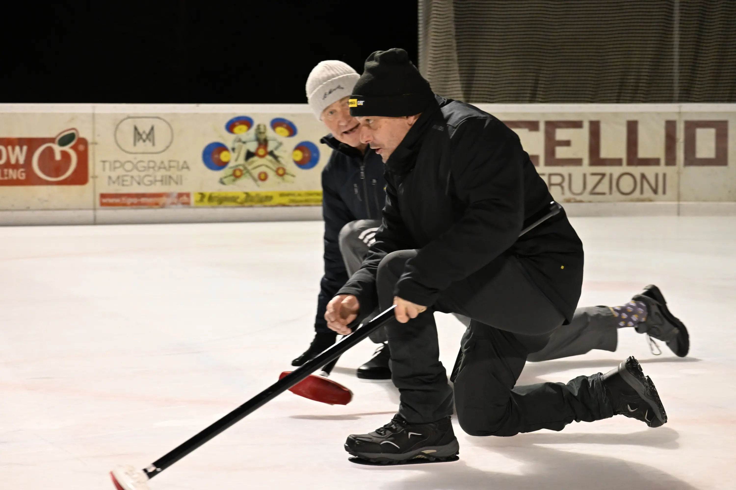 Due persone inginocchiate sul ghiaccio mentre giocano a curling, concentrandosi sul posizionamento della pietra. Indossano abbigliamento invernale e guanti.