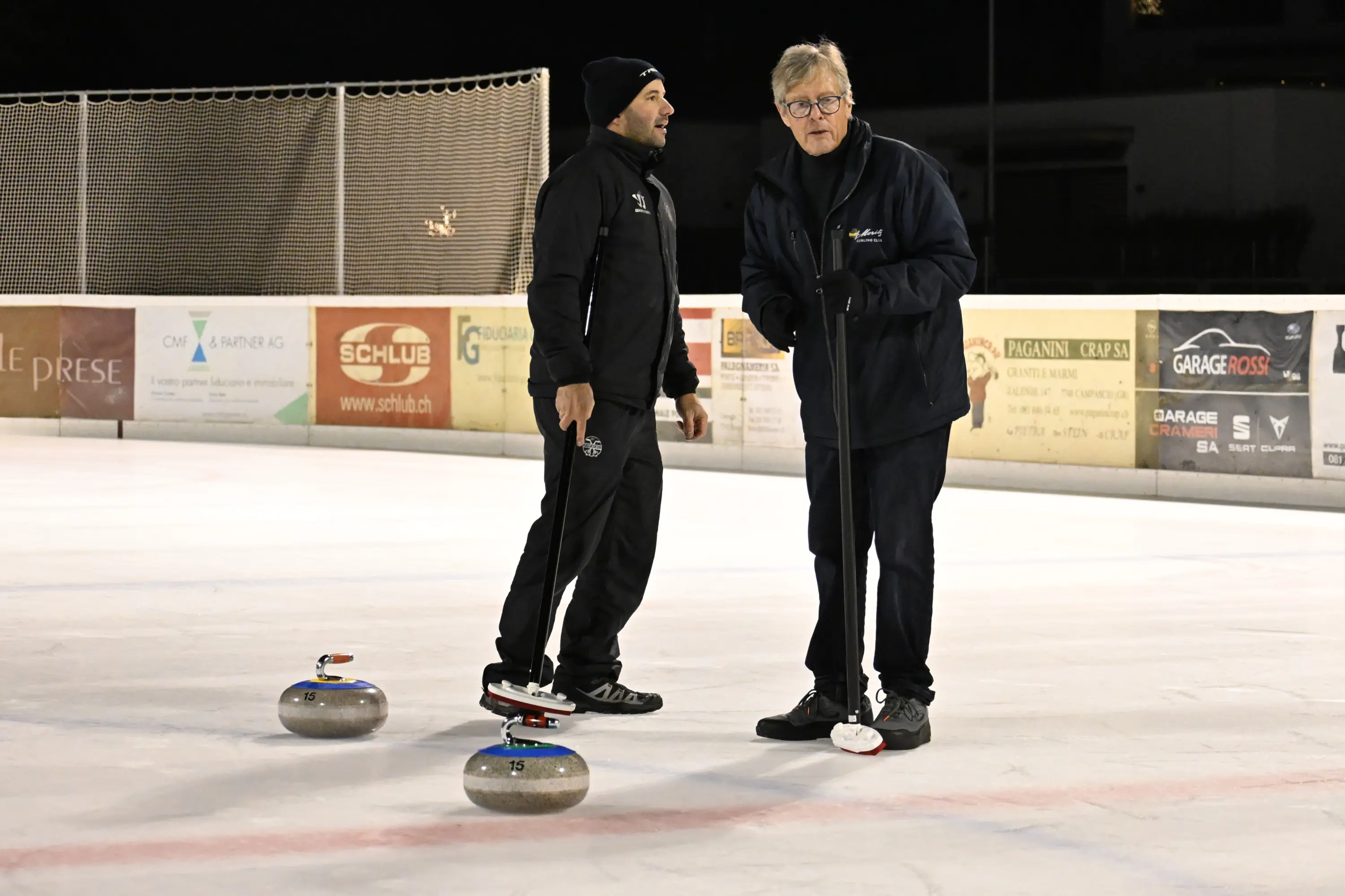 Due uomini si trovano su un campo di curling, uno è in piedi accanto a una pietra da curling, mentre l'altro usa una scopa. Sullo sfondo si intraved