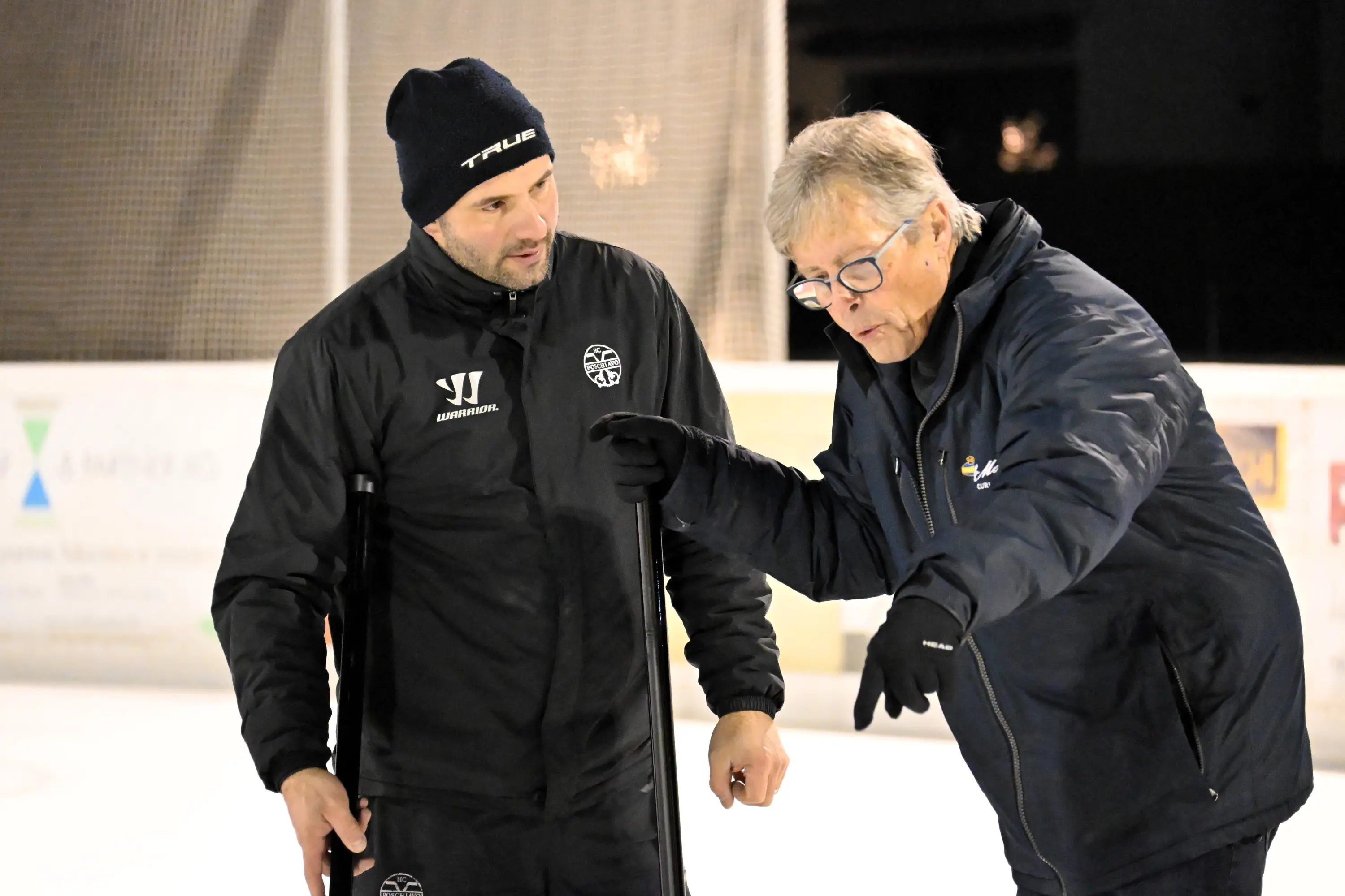 Due uomini discutono su un campo da hockey sul ghiaccio, uno indossa un berretto e giacca nera, l'altro una giacca blu. Sembri concentrati mentre parl