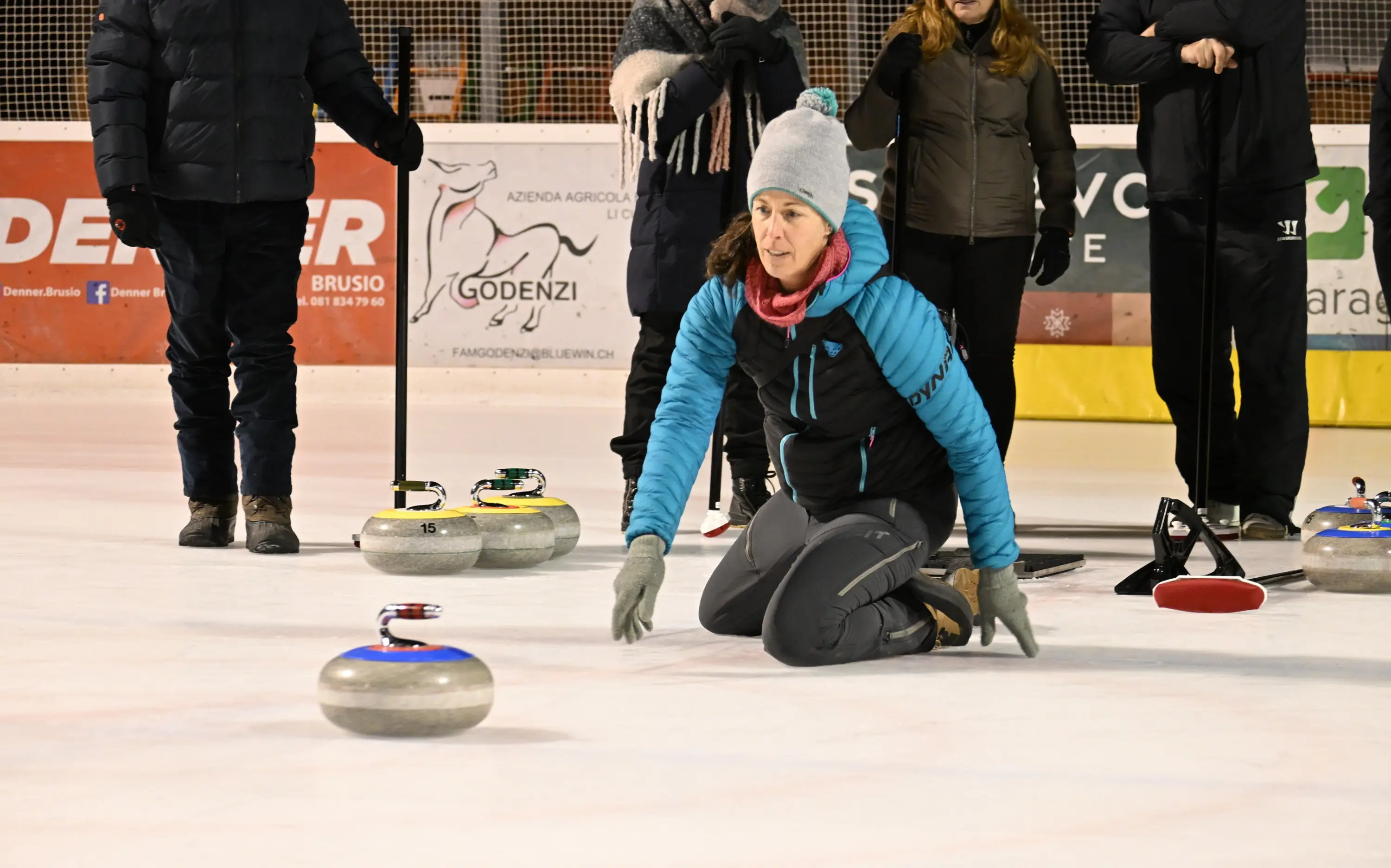 Una donna in posizione inginocchiata sulla pista di curling, mentre lancia una pietra. Indossa un abbigliamento sportivo e un cappello grigio. Sullo sf