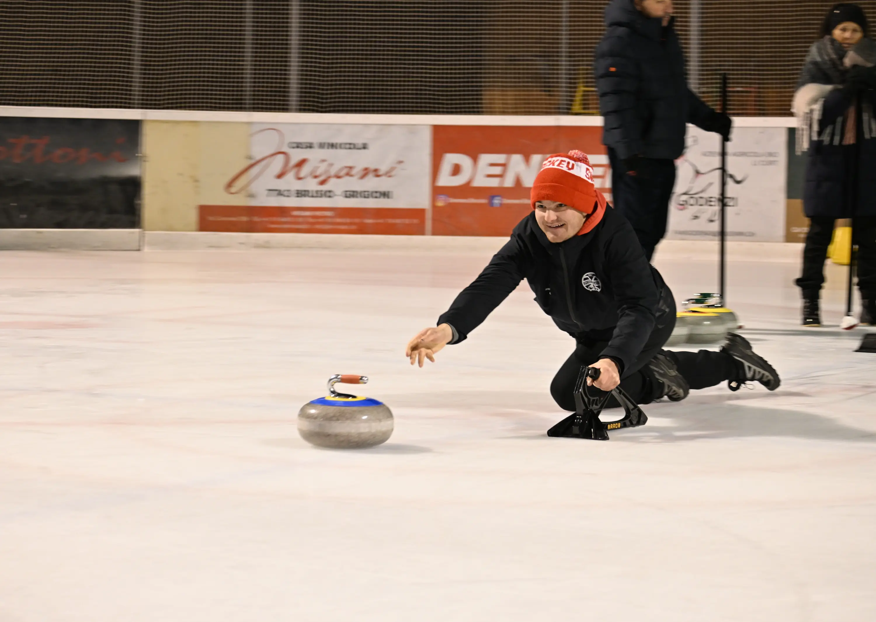 Un uomo in tuta nera e cappello rosso si proietta in avanti mentre lancia un disco di curling su una pista di ghiaccio. Sullo sfondo si intravedono altre