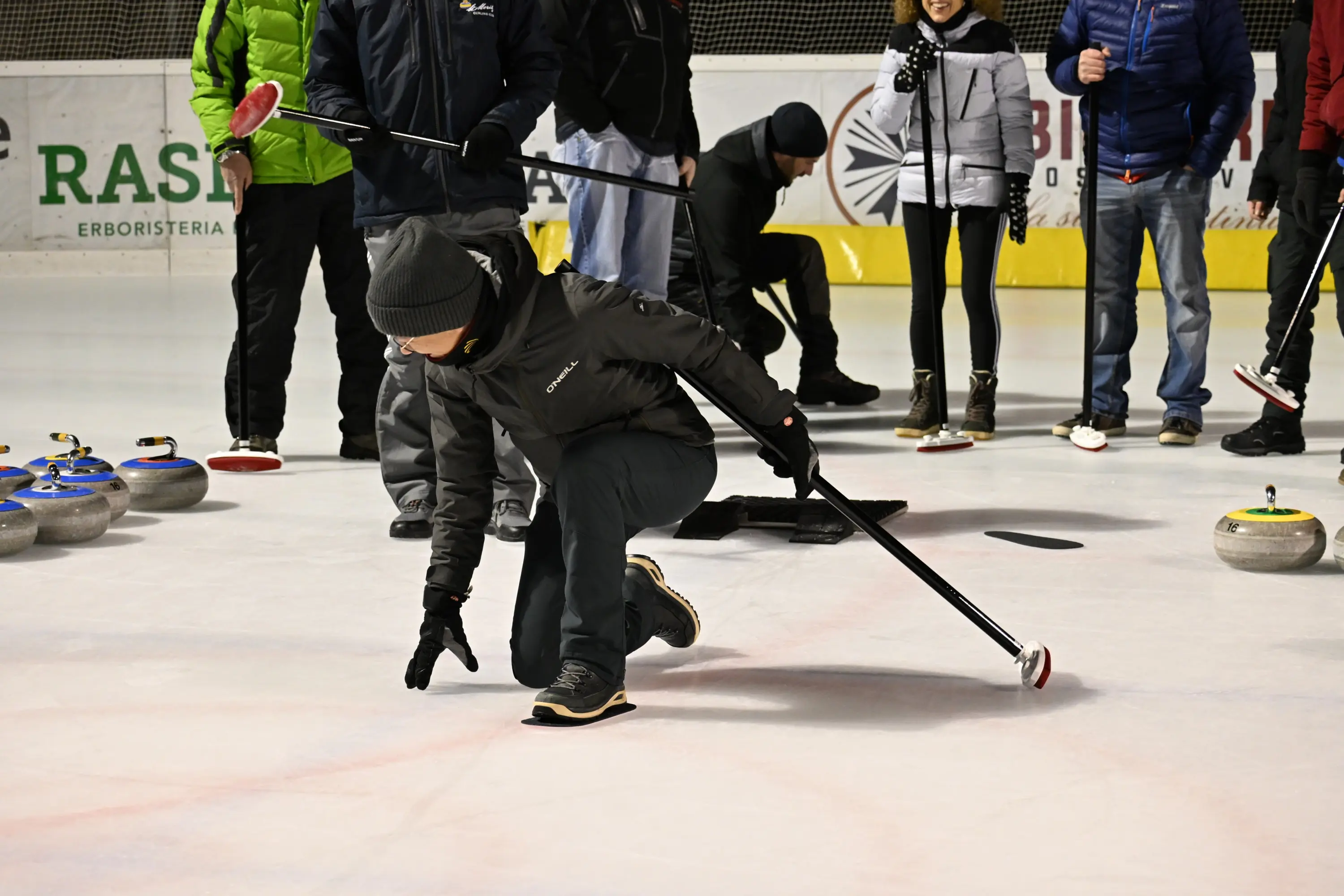 Una persona si inginocchia sul ghiaccio mentre gioca a curling, lanciando una pietra. Altre persone osservano in background, indossando abbigliamento invernale