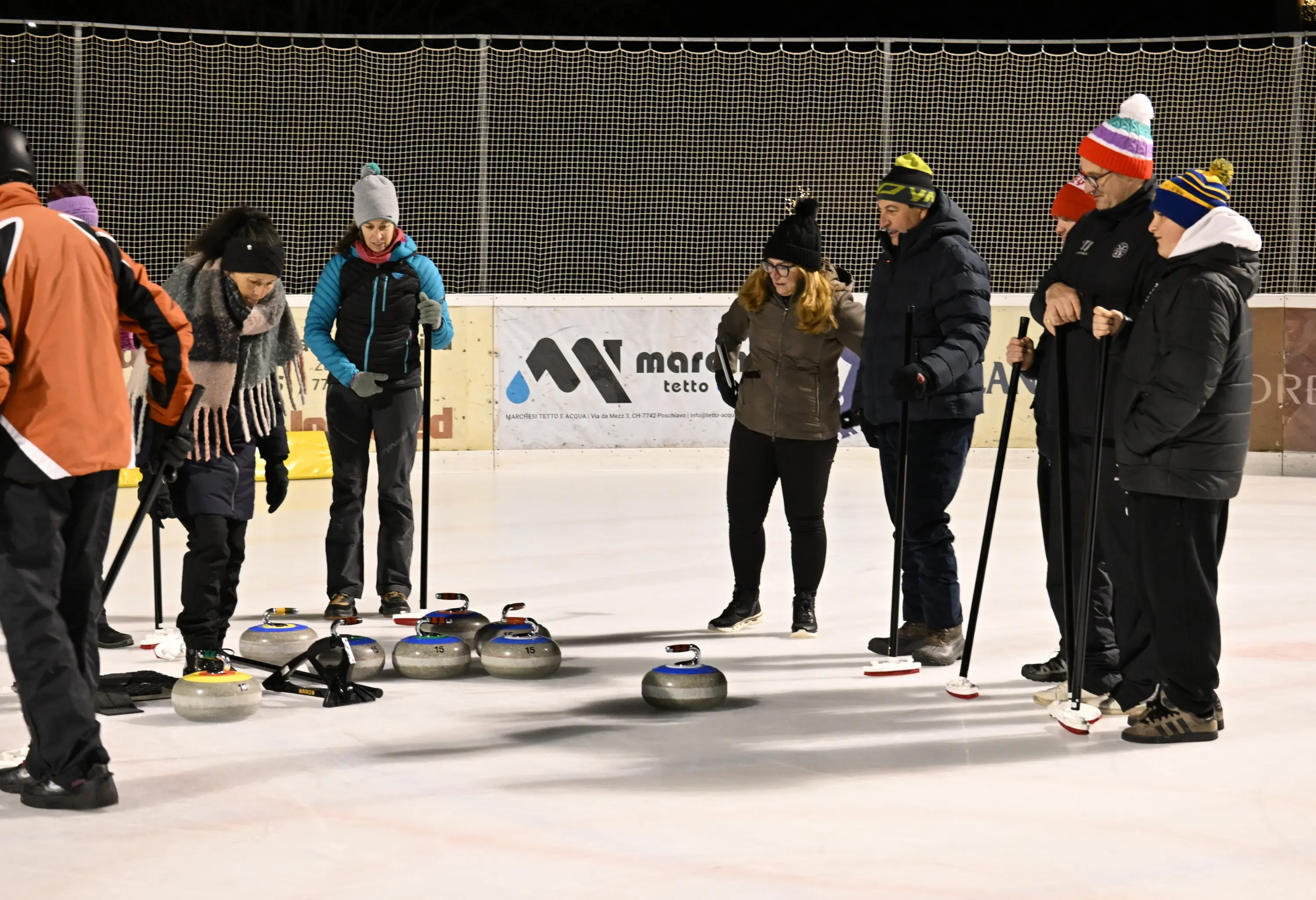 Gruppo di persone su una superficie di ghiaccio, intente a giocare a curling. Alcuni tengono delle stecche, mentre altri si piegano per osservare le pietre