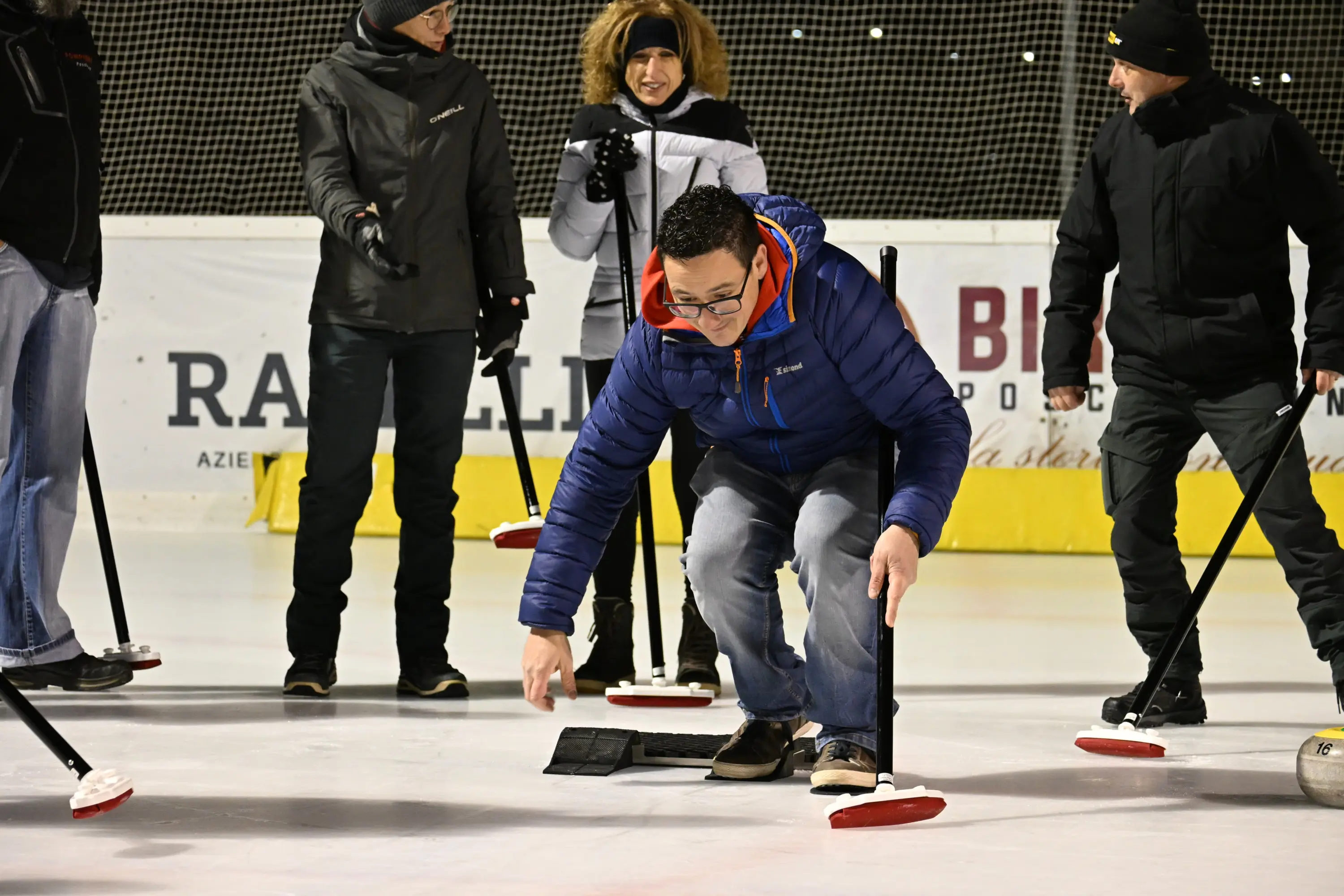 Un gruppo di persone si trova su un campo di curling, con un uomo che si piega in avanti per lanciare una pietra. Gli altri partecipano attivamente, alcuni tengono le