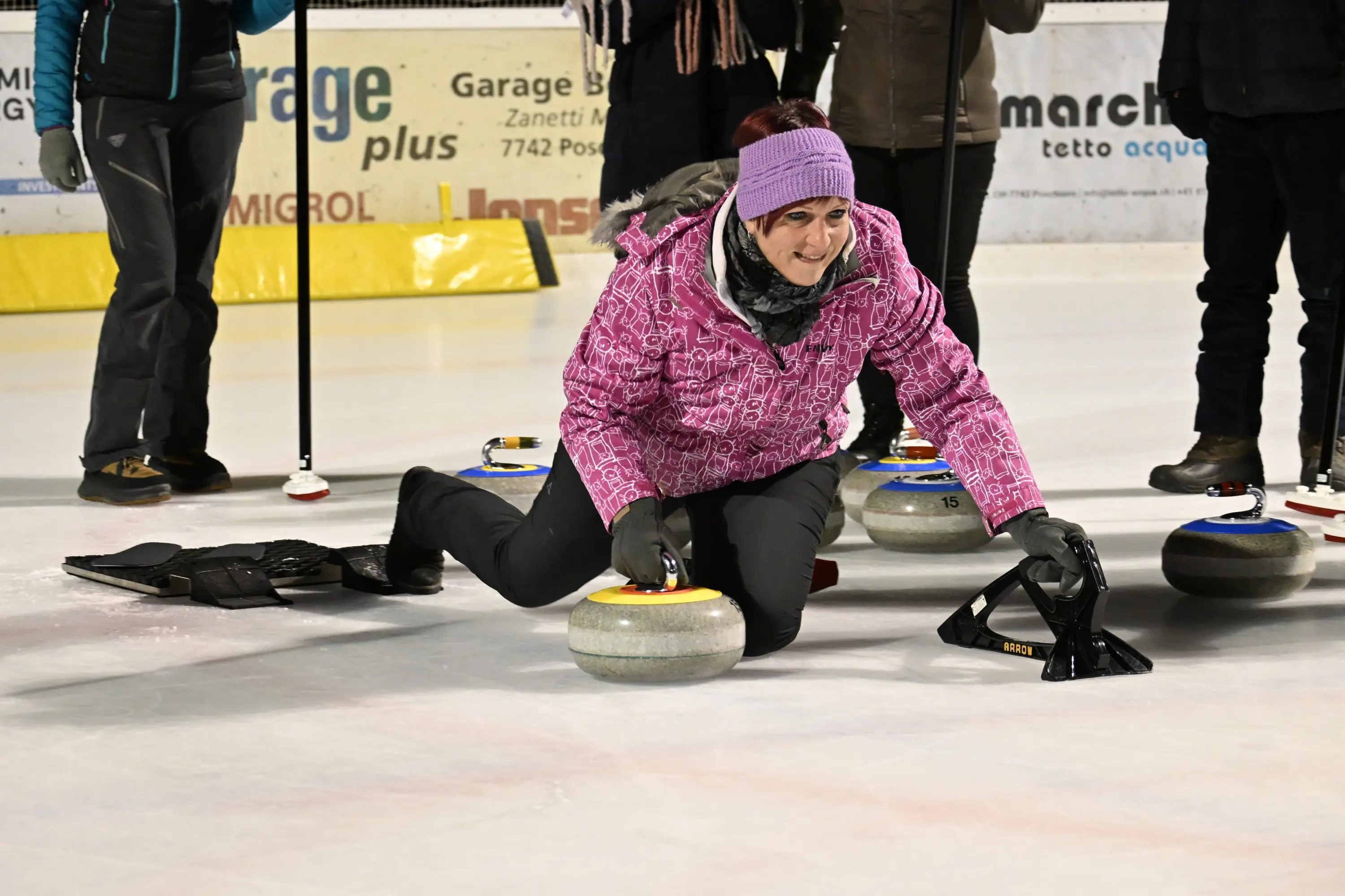 Una donna in giacca rosa si china mentre lancia una pietra da curling su un campo di ghiaccio. Indossa guanti neri e un berretto, e ci sono altre persone e