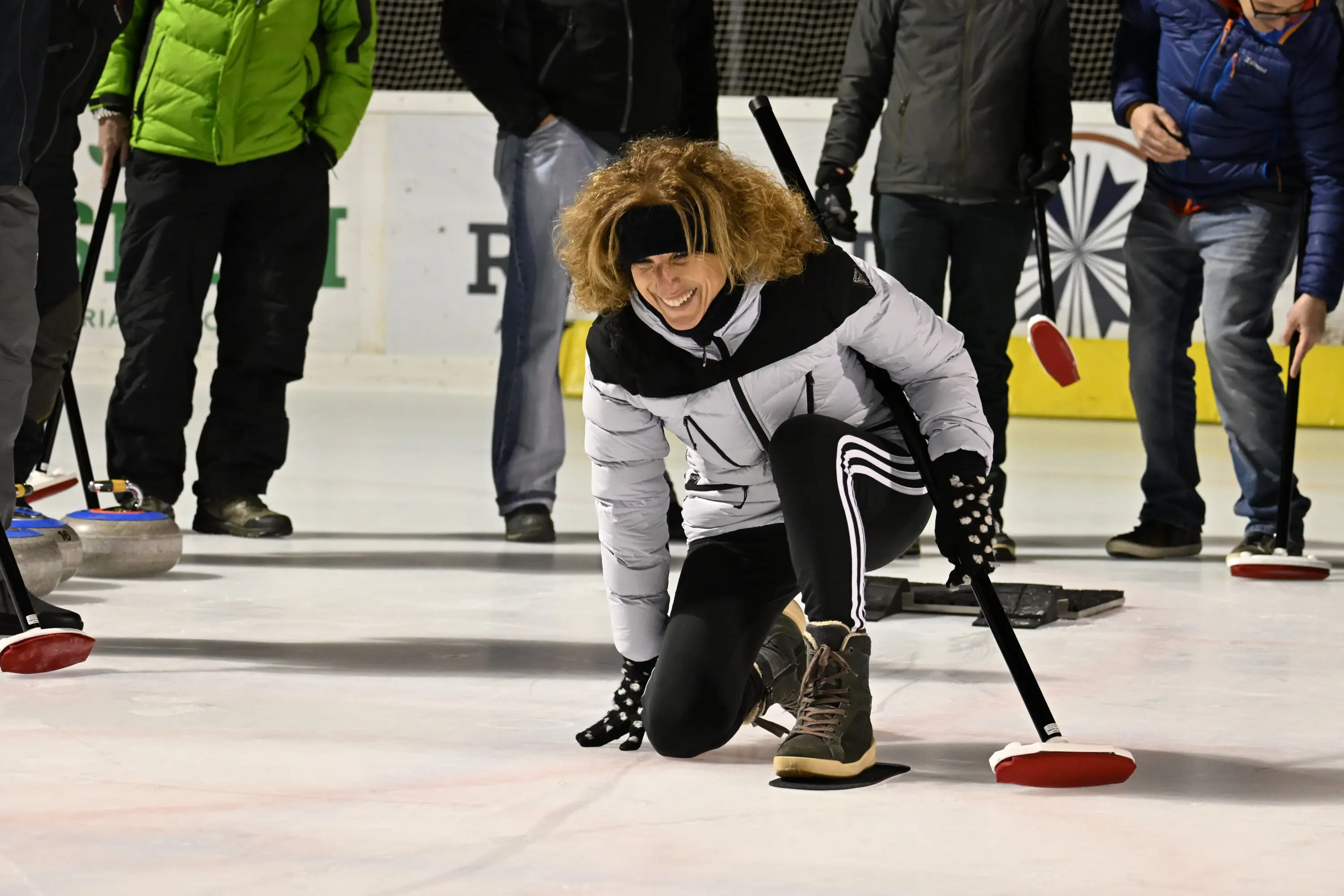 Una donna con capelli ricci e biondi, vestita con un piumino bianco e pantaloni neri, si piega in avanti mentre pratica curling su una pista di ghiaccio.