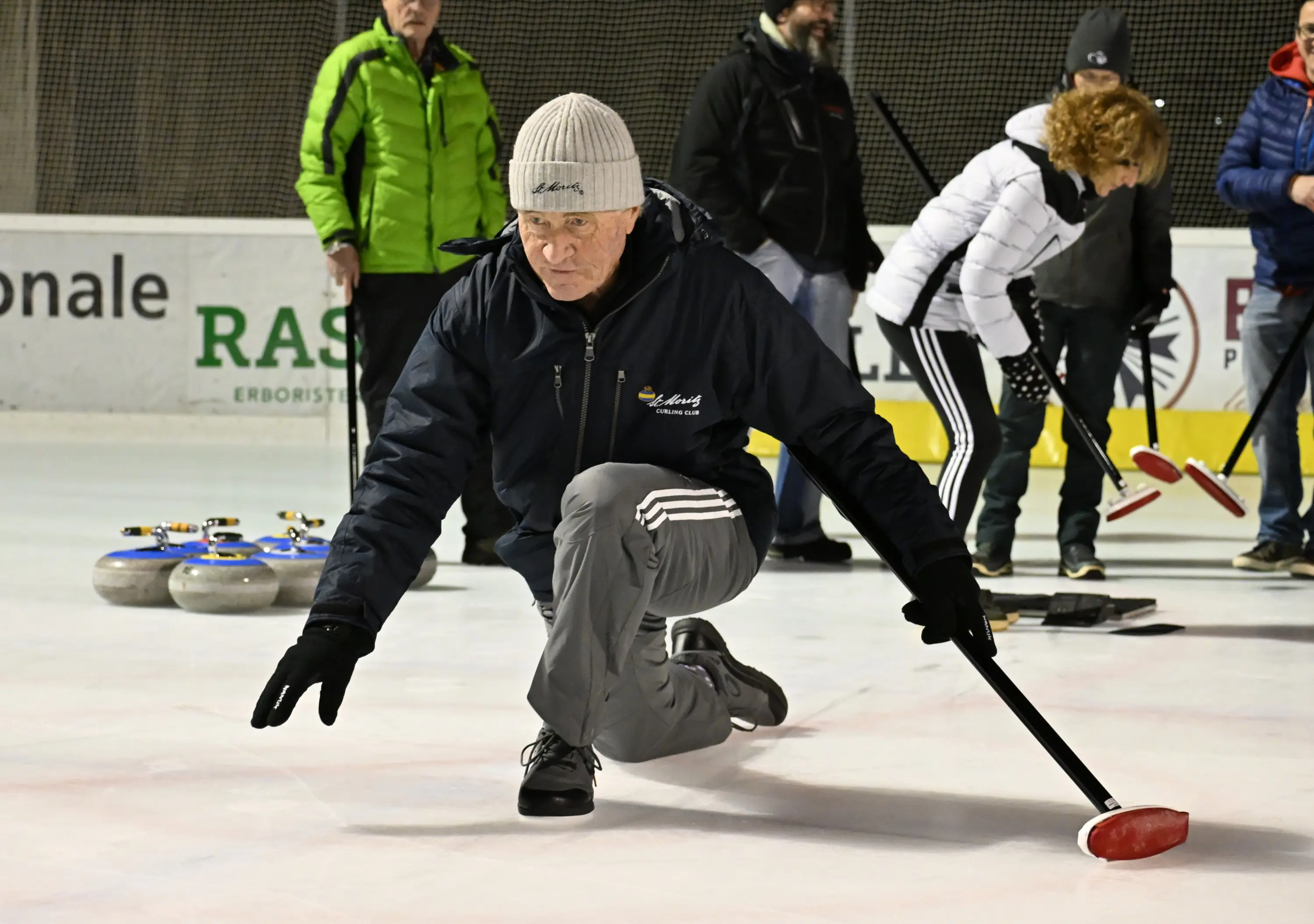 Uomo anziano in giacca blu e berretto grigio, mentre si prepara a lanciare una pietra da curling sul ghiaccio. Sullo sfondo, altre persone sono coinvol