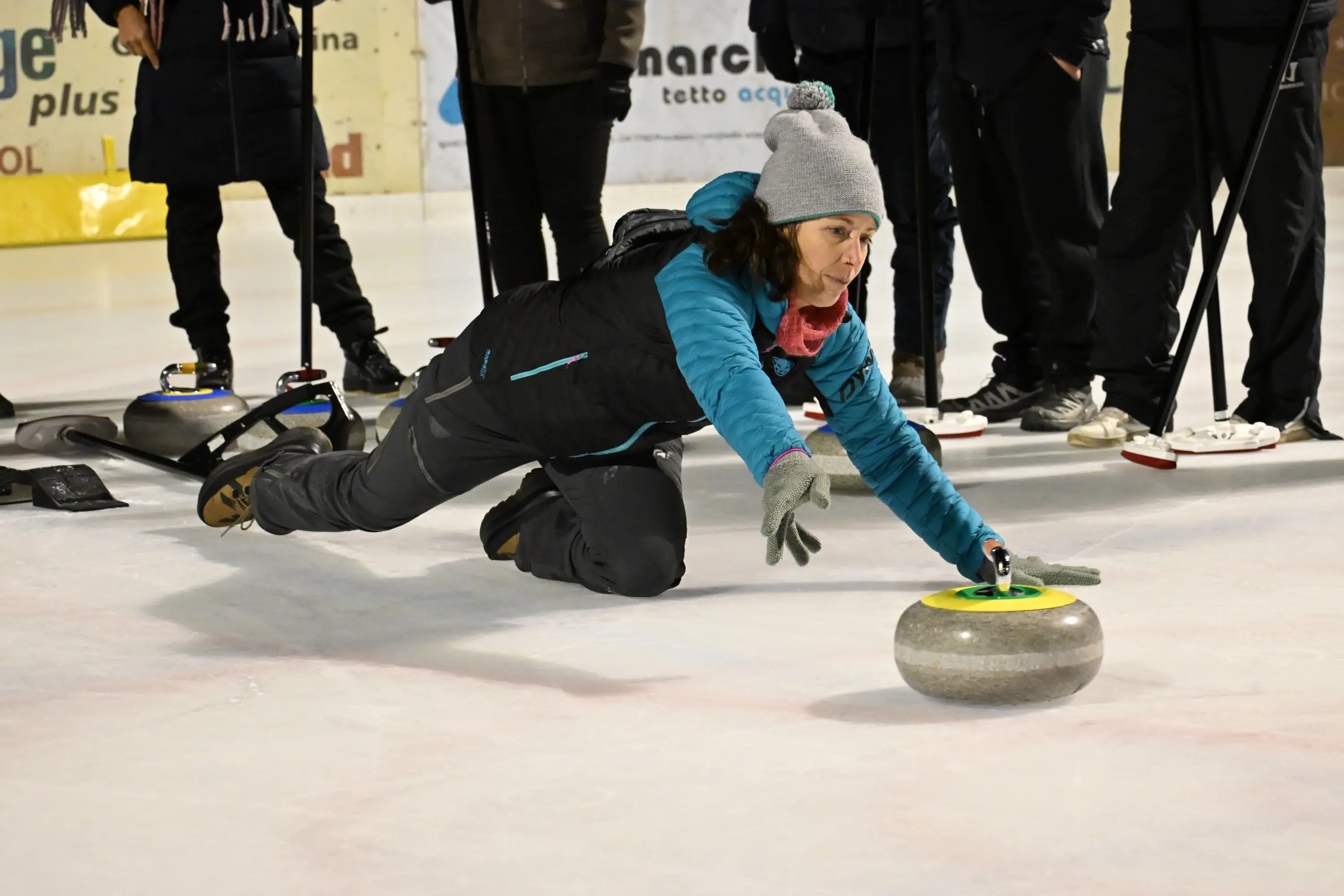 Una persona si sta lanciando una pietra da curling sul ghiaccio, in una posizione bassa e concentrata, indossando un giaccone e un berretto. Sullo sf