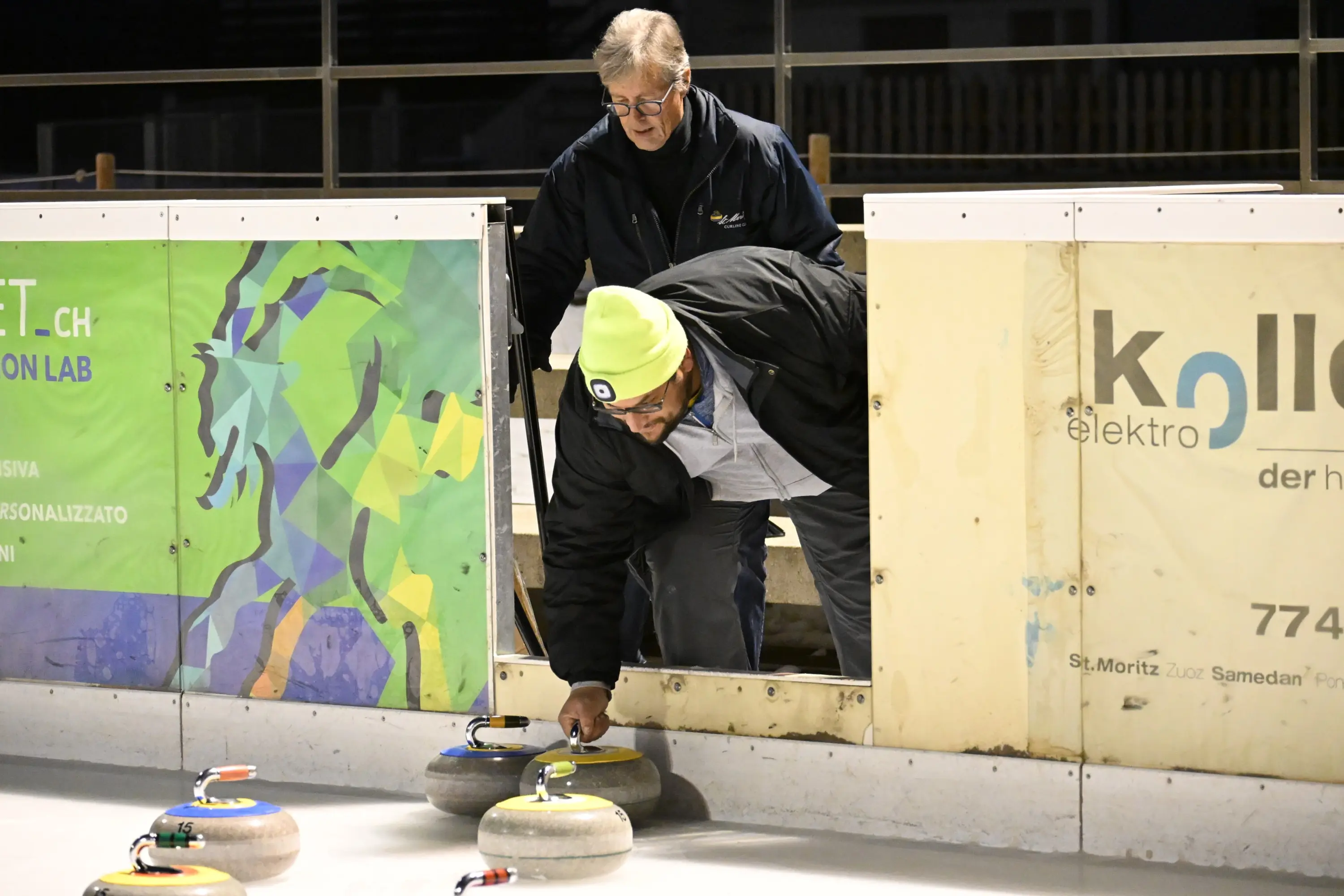 Due persone si trovano vicino al bordo di un campo da curling. Un uomo indossa un cappello giallo e sta sistemando una pietra, mentre l'altro lo osserva attent